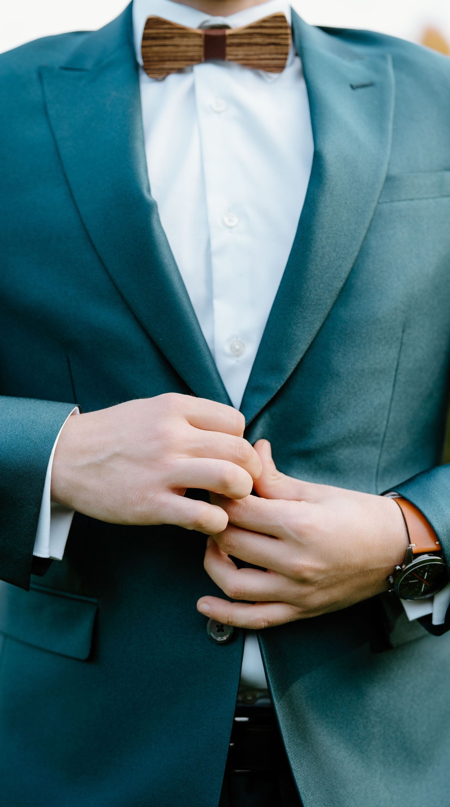 Close-up of hands adjusting a teal suit jacket with wooden bow tie and white dress shirt.