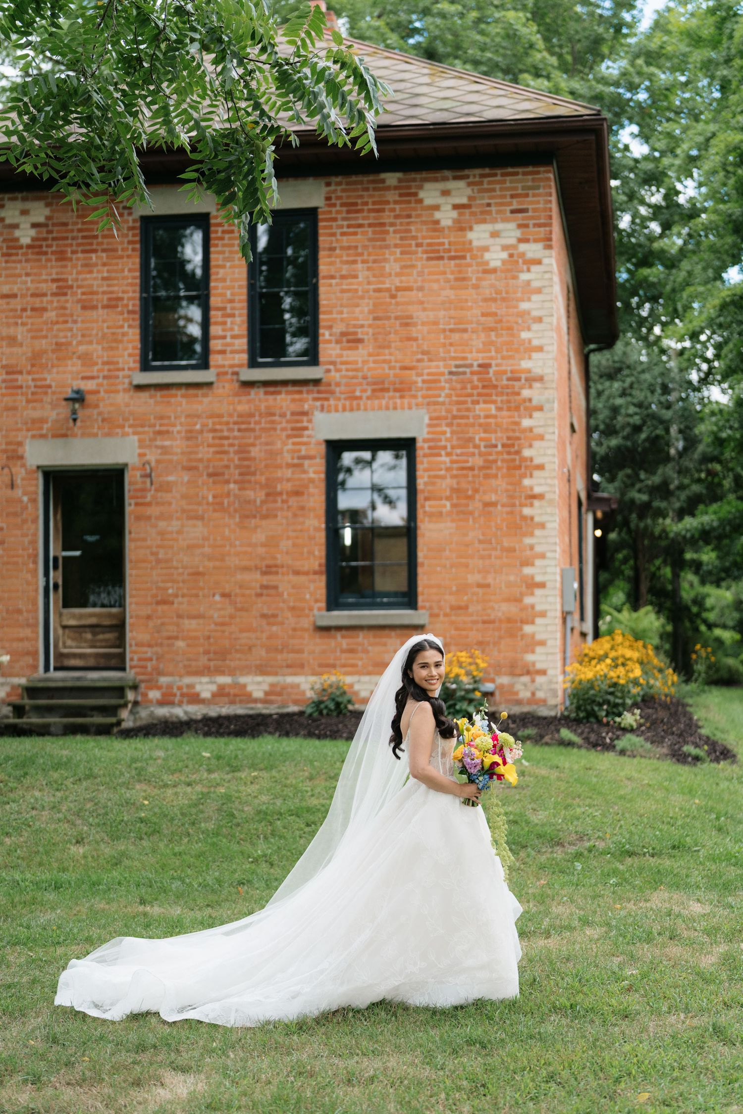 A wedding scene at a rustic brick house with a flowing white dress trailing on the green lawn.