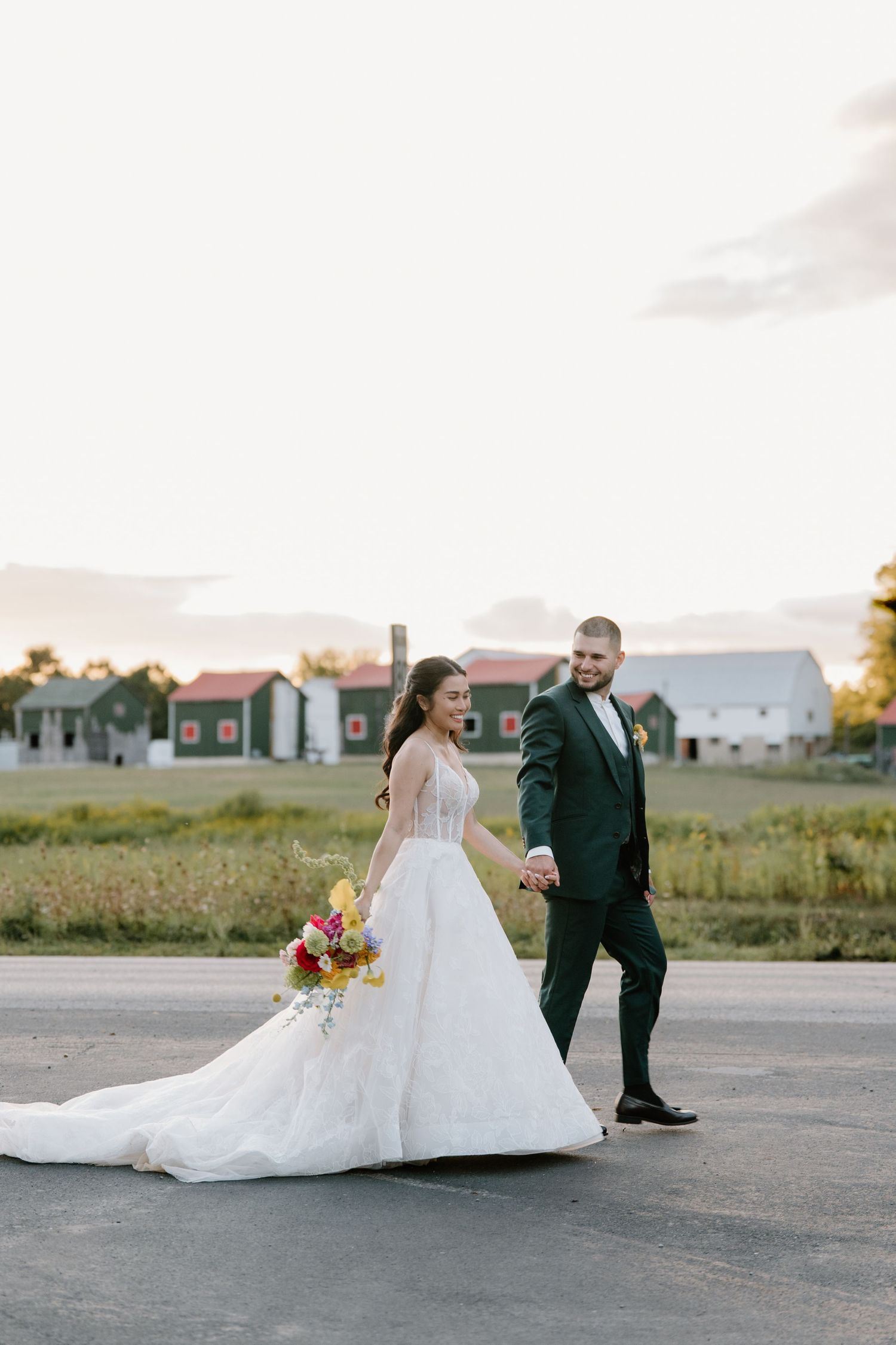 A couple in wedding attire poses artistically on a paved surface with buildings and fields in the background at sunset.
