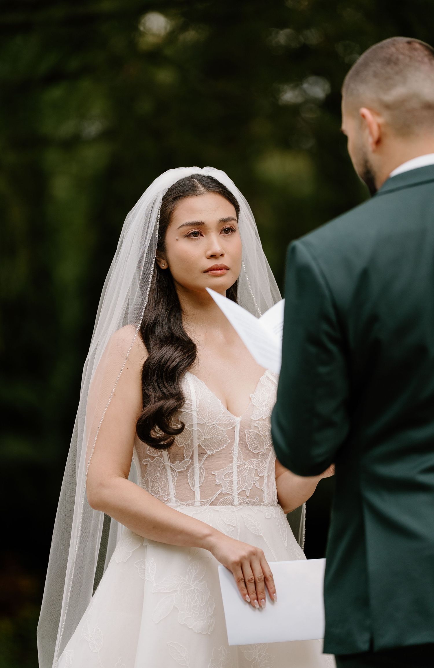 A bride in a white corset wedding dress wipes away tears during an emotional ceremony moment.