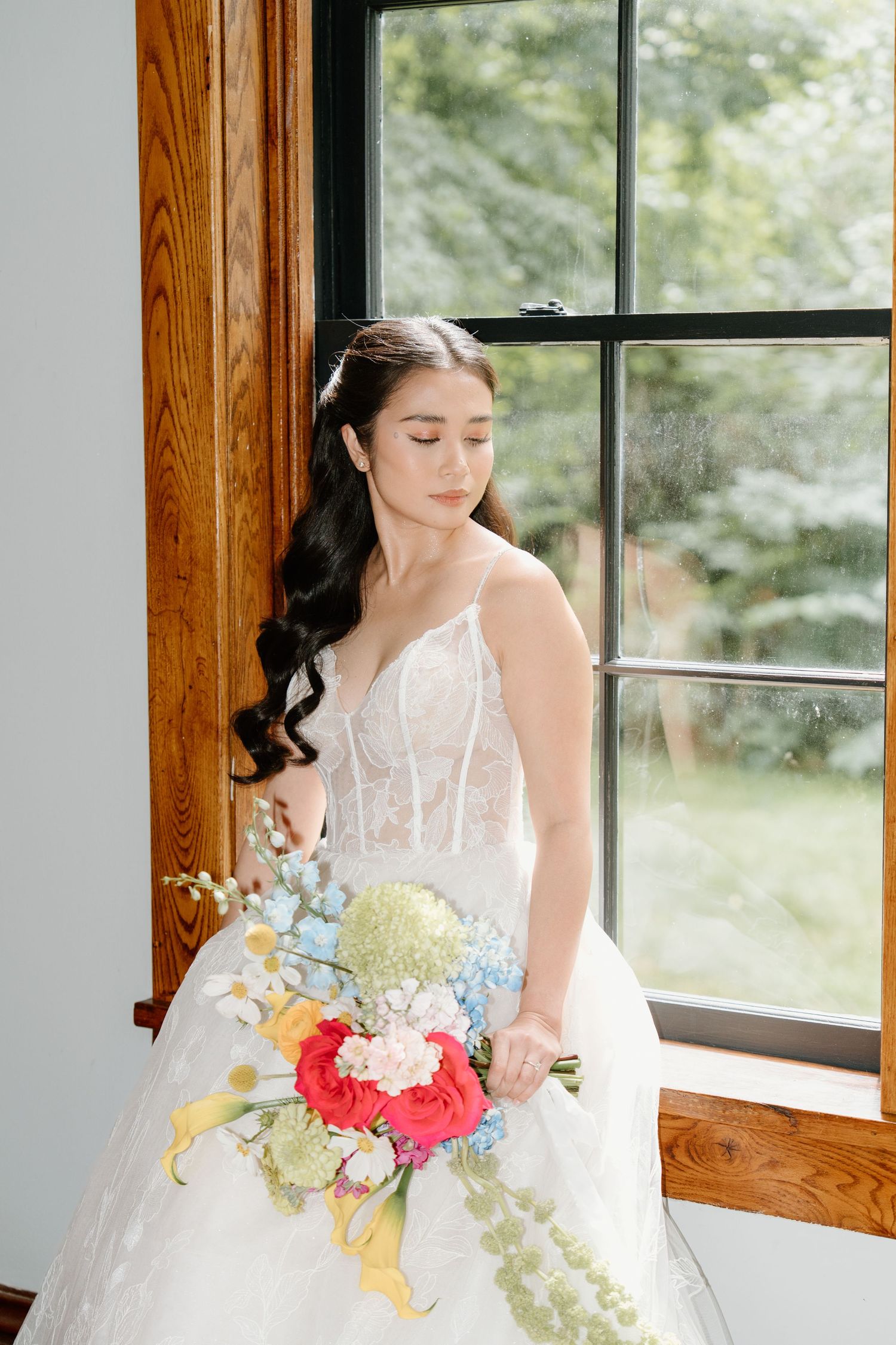 Bride in white ballgown holds colorful bouquet featuring pink and blue flowers near sunlit window.