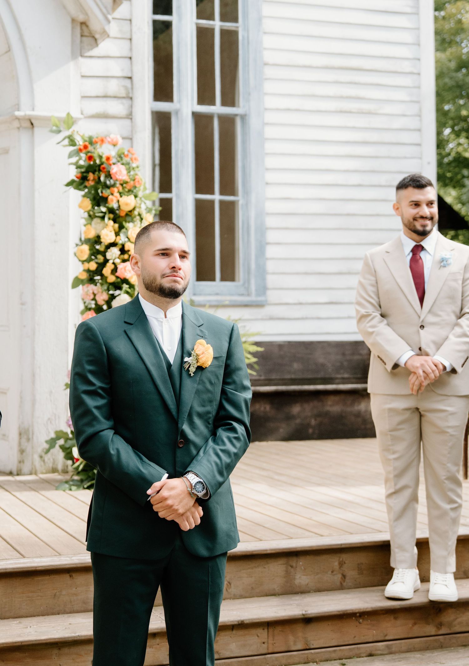 Wedding party members in green and beige suits stand on a white wooden deck of a historic chapel.