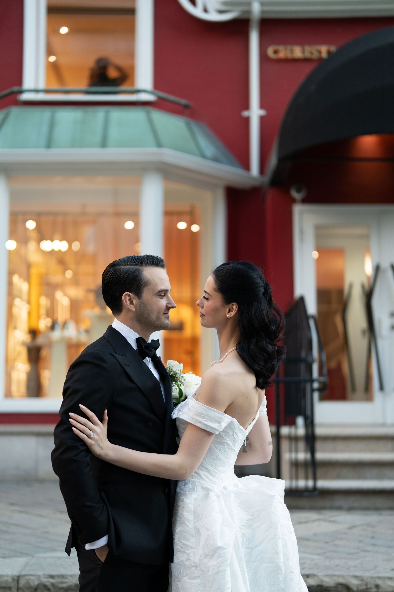 A romantic wedding couple embraces on a cobblestone street in front of a red building with illuminated storefronts.