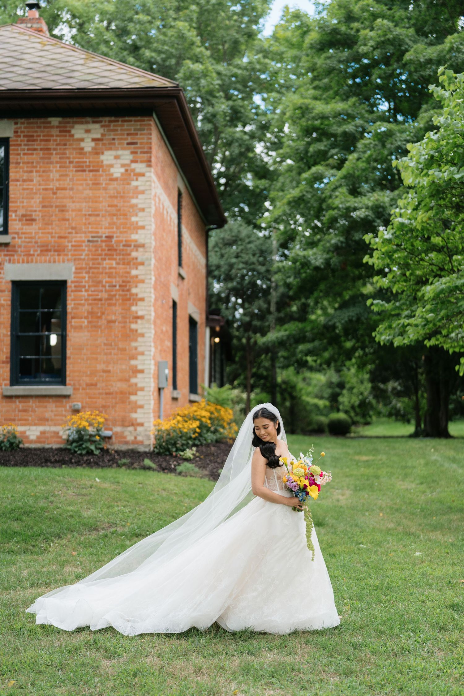 A flowing white wedding dress cascades across green grass near a brick building on a sunny summer day.