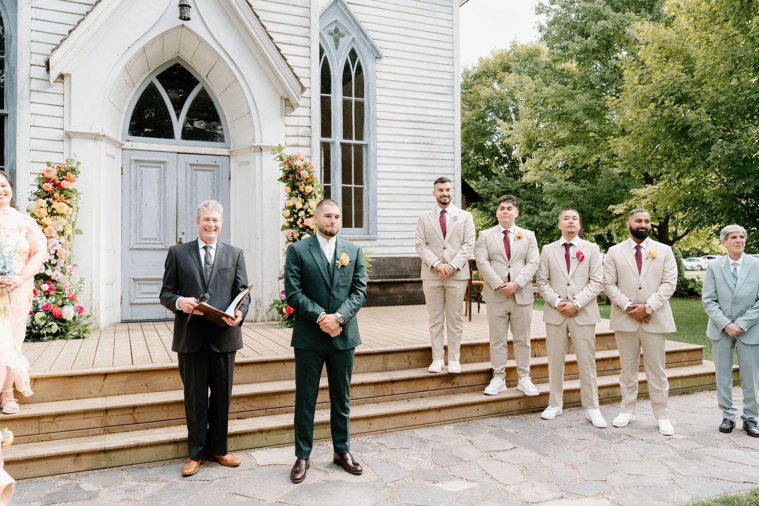 Wedding party stands on the steps of a white chapel with Gothic arched doorways and floral decorations.
