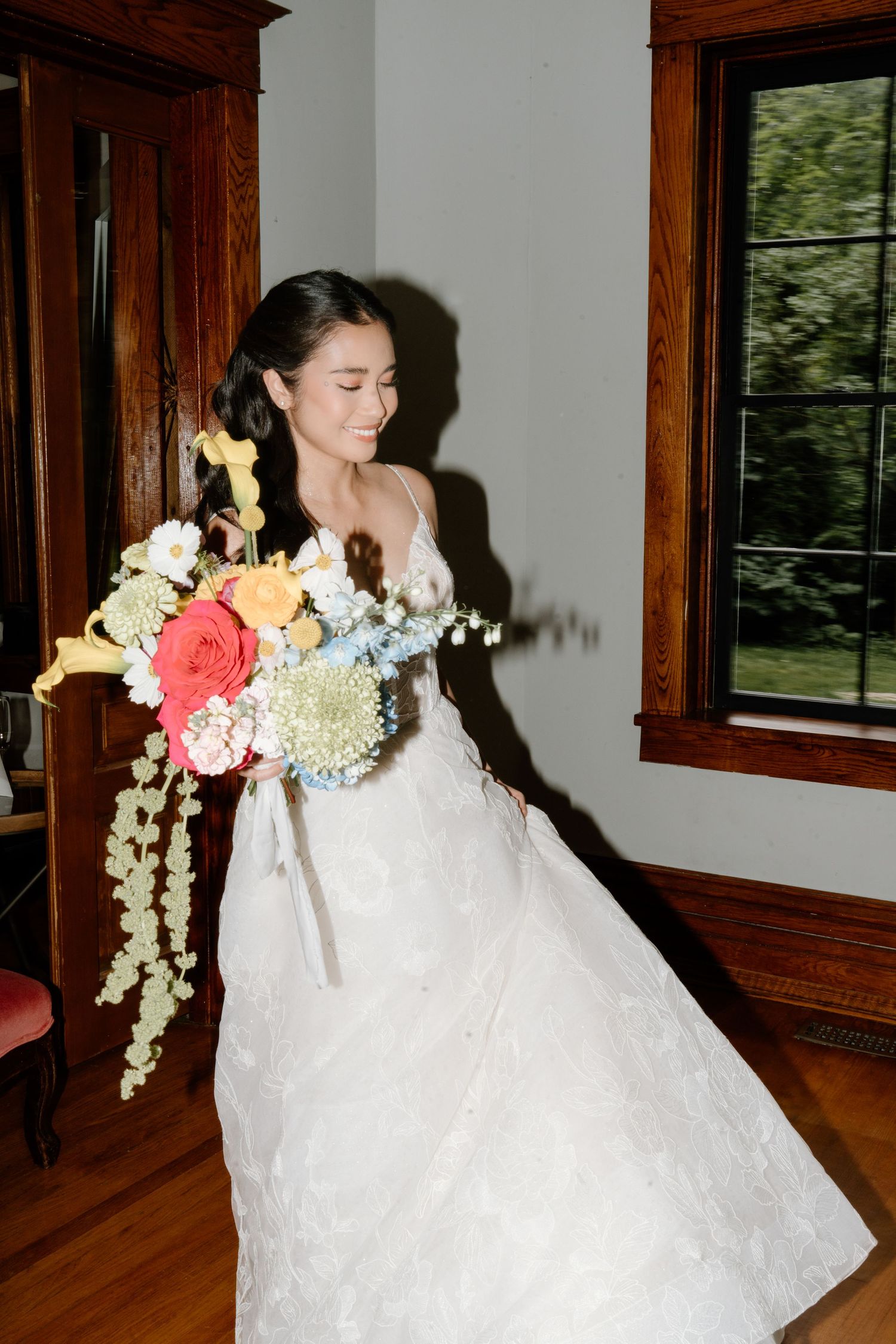 A bride in a white lace wedding dress holds a vibrant bouquet of coral and white flowers in a wood-trimmed room.