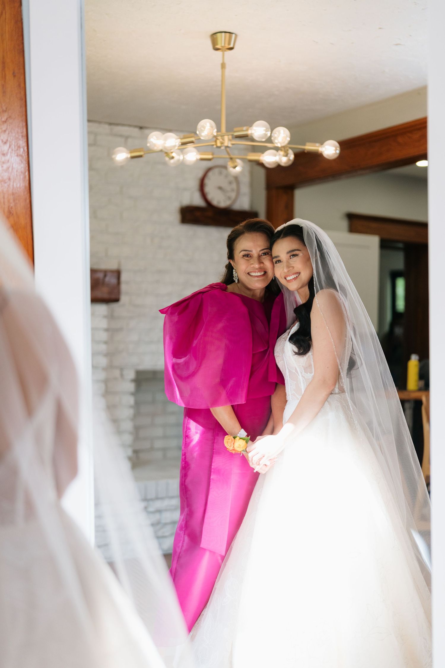 A joyful wedding scene with a bride in white and companion in vibrant pink sharing laughter in an indoor setting.