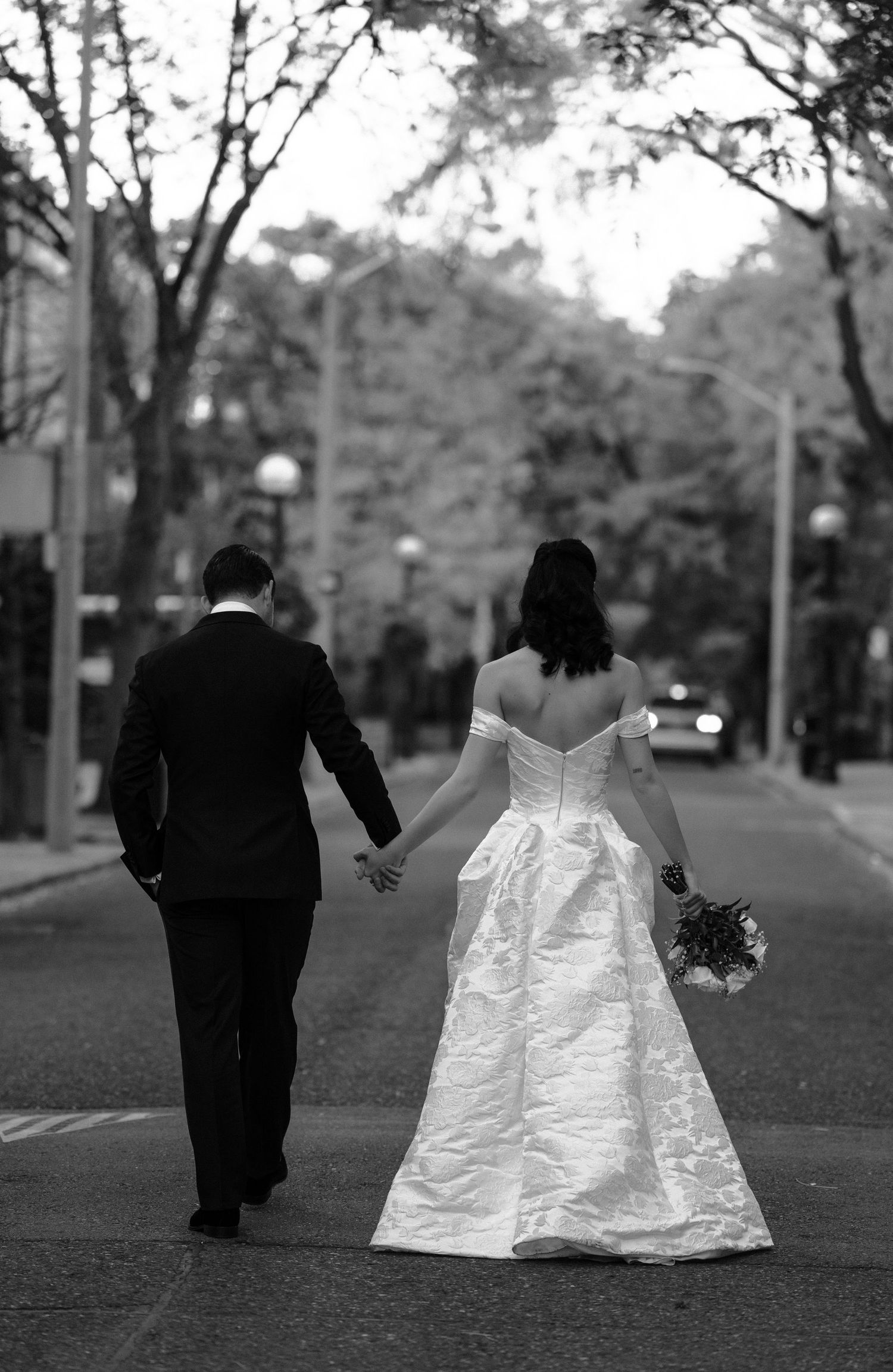 A couple in wedding attire walking hand in hand down a tree-lined street in black and white photography.