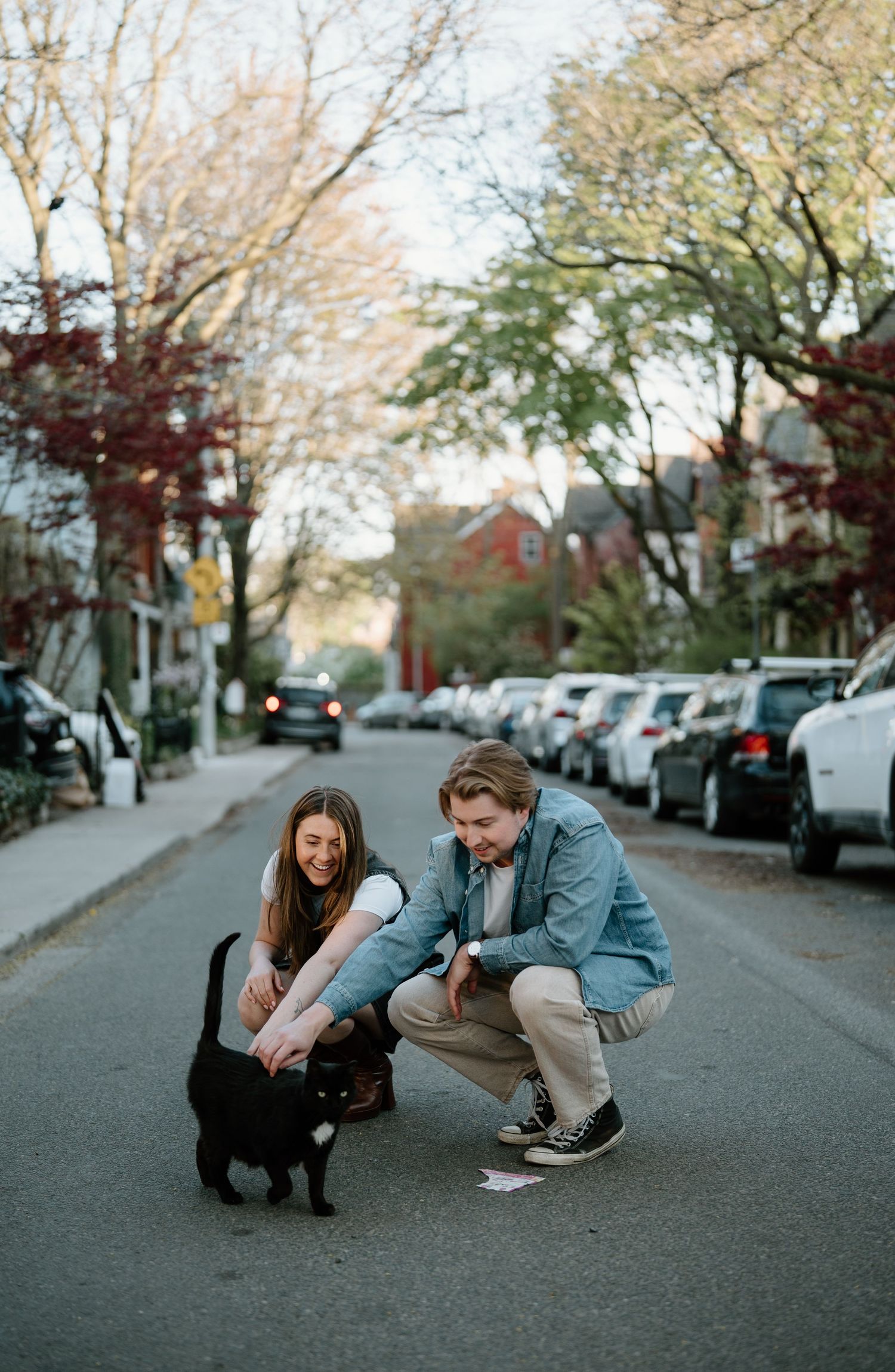 A couple crouches down to pet a black cat on a quiet residential street lined with parked cars and autumn trees.