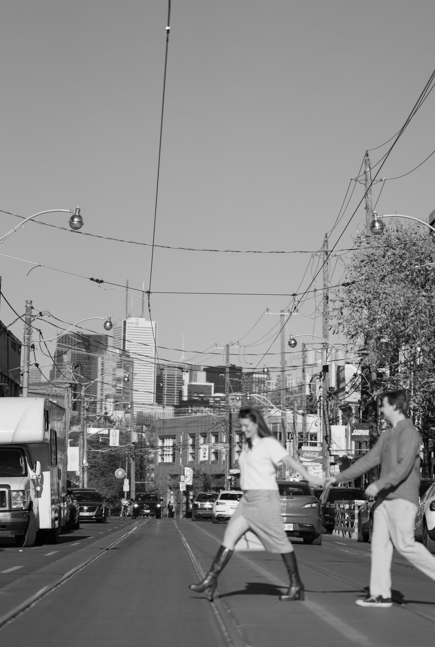 Black and white urban street scene in a bustling commercial district with trolley lines overhead and pedestrians crossing.