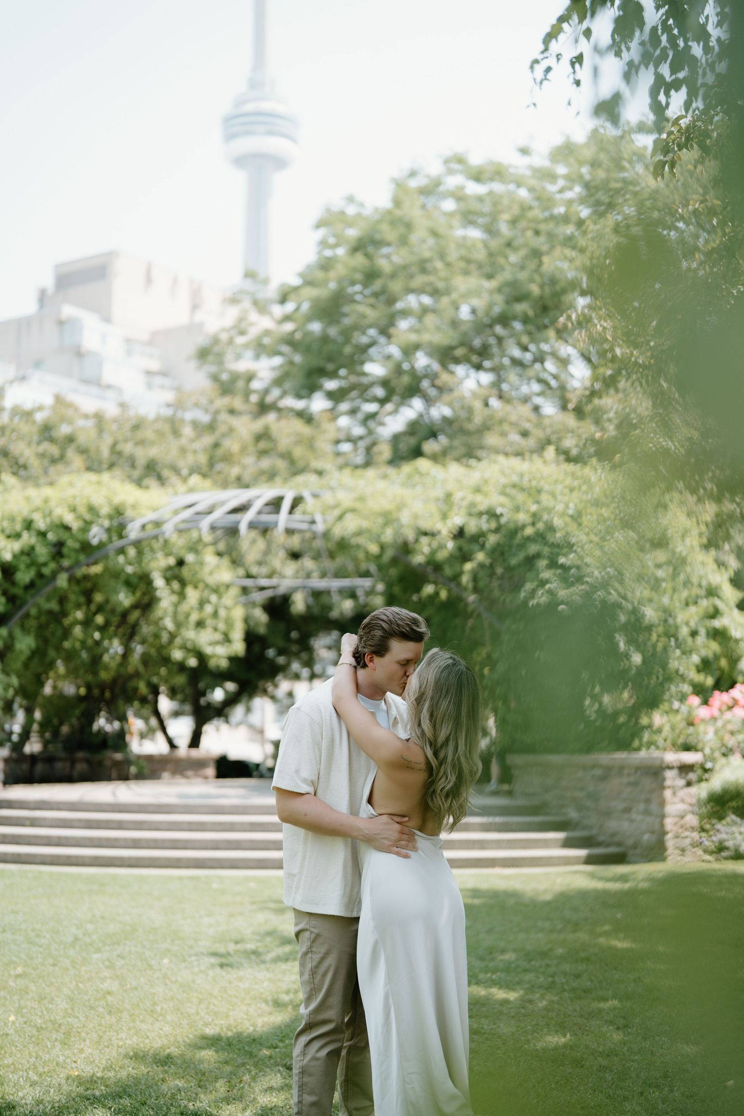 A romantic couple embraces in a lush garden park with the CN Tower visible in the Toronto skyline background.