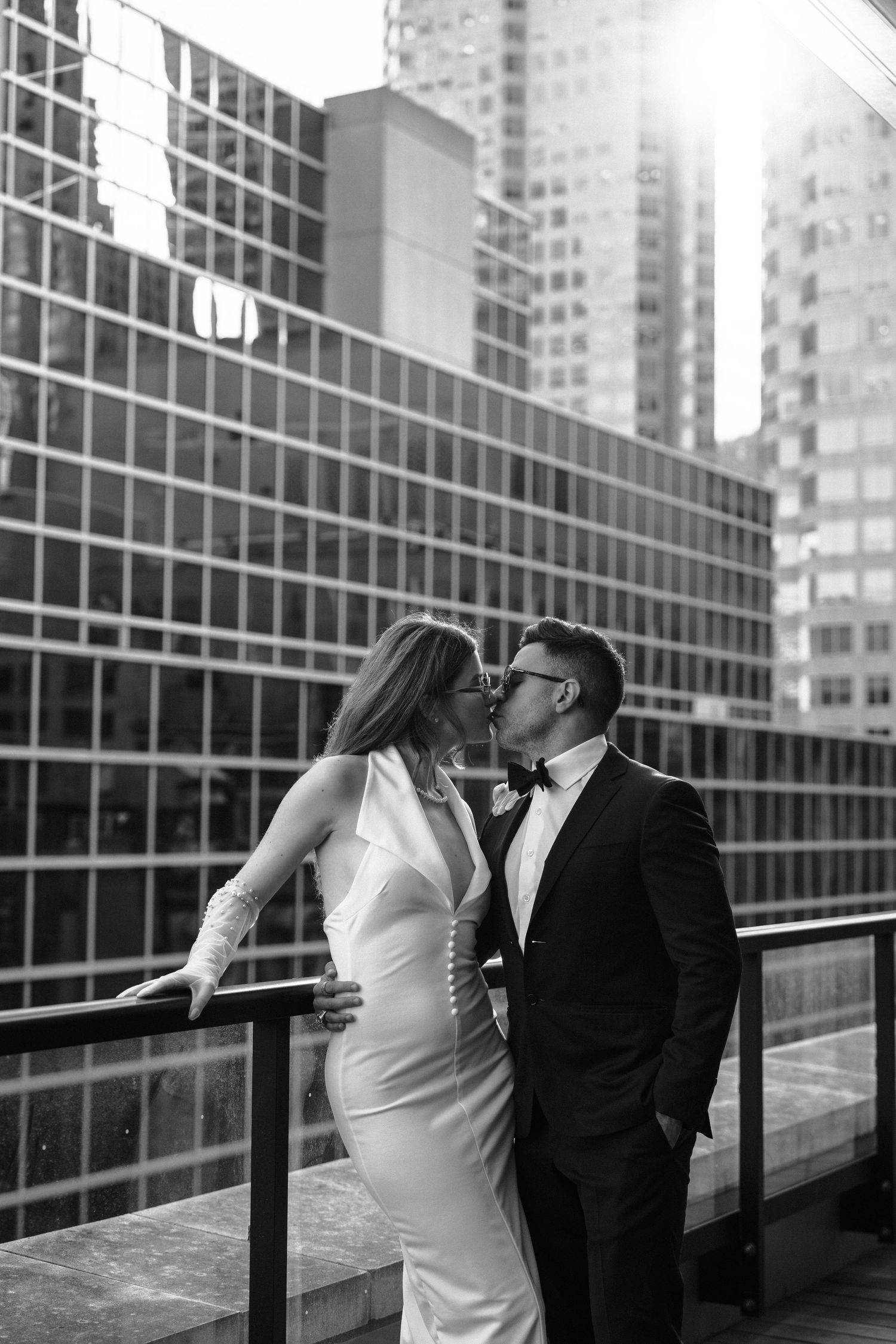 A couple shares a romantic kiss on a rooftop balcony with modern city buildings creating a dramatic urban backdrop.