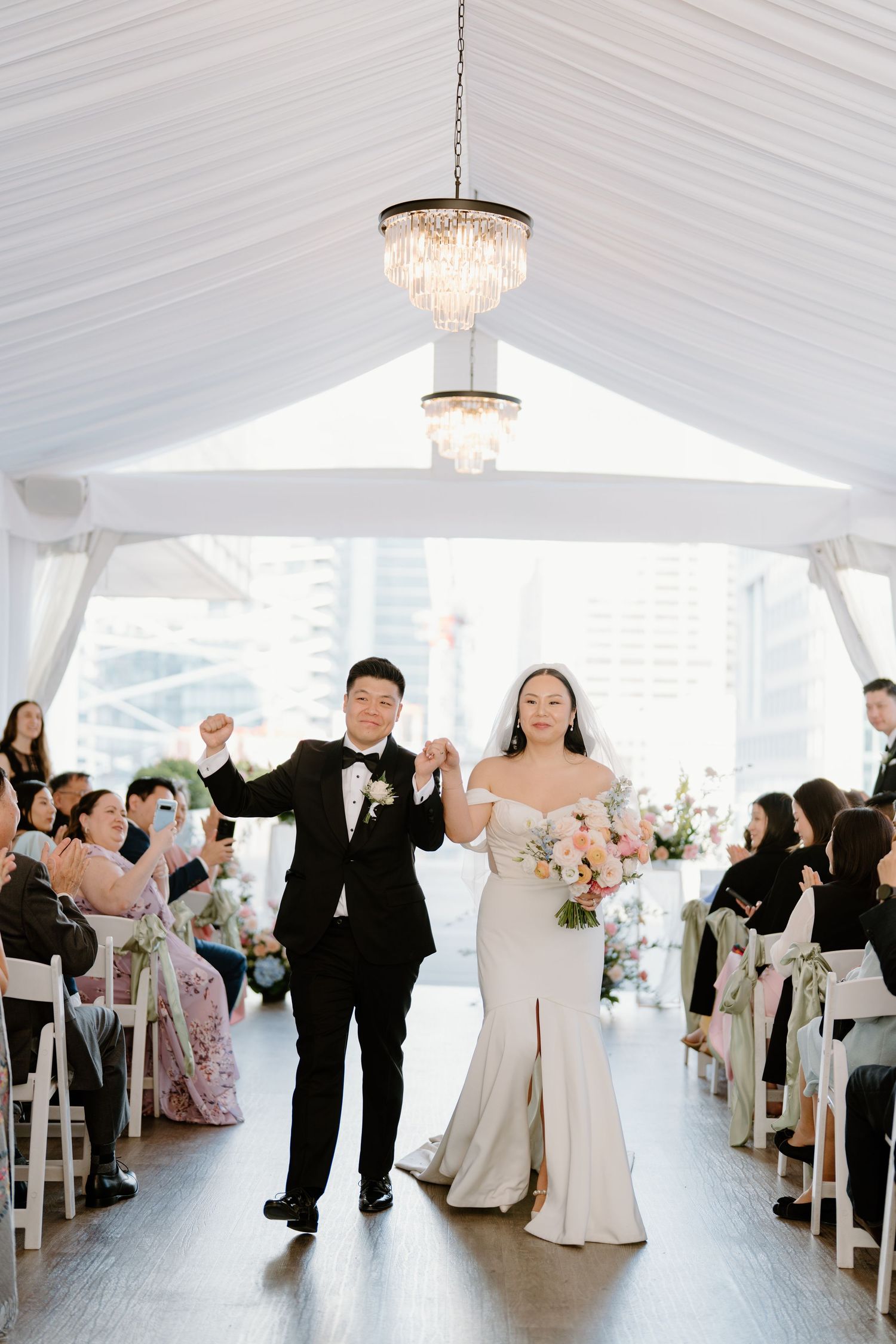 Newlyweds celebrate joyfully walking down aisle under crystal chandeliers in white tent wedding ceremony.