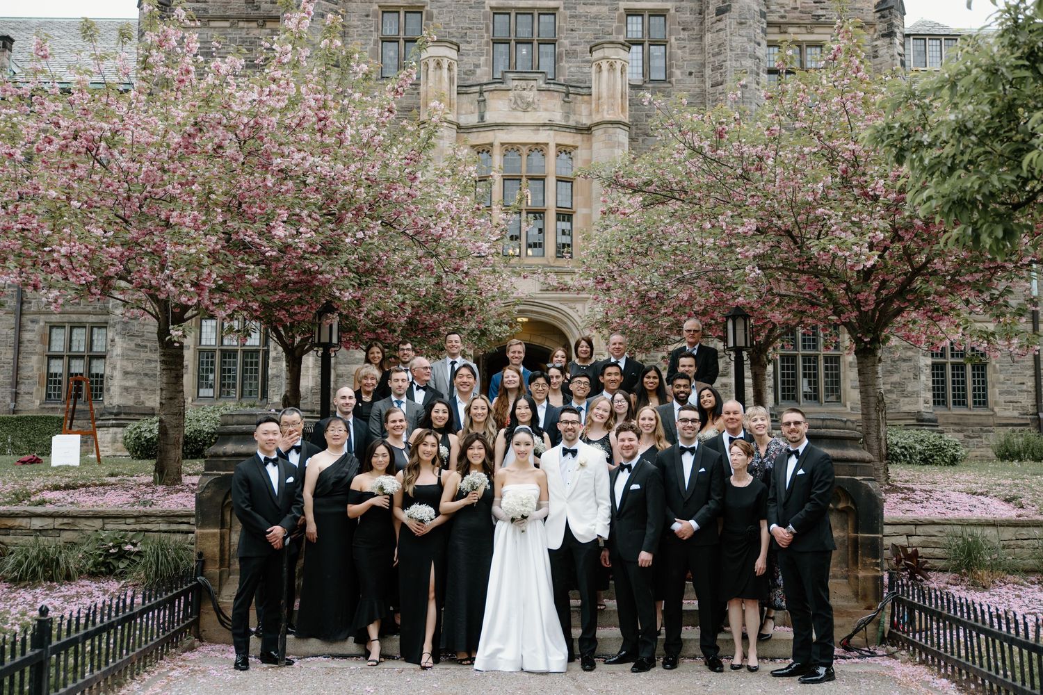 A wedding party poses together in front of a gothic stone building with spring cherry blossoms blooming.