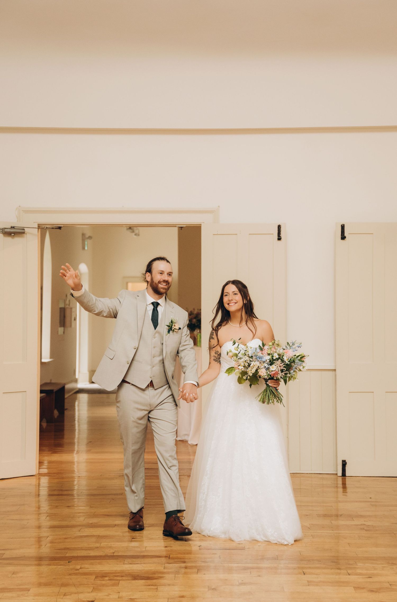 A couple in wedding attire dances playfully in a bright indoor venue with hardwood floors and white walls.