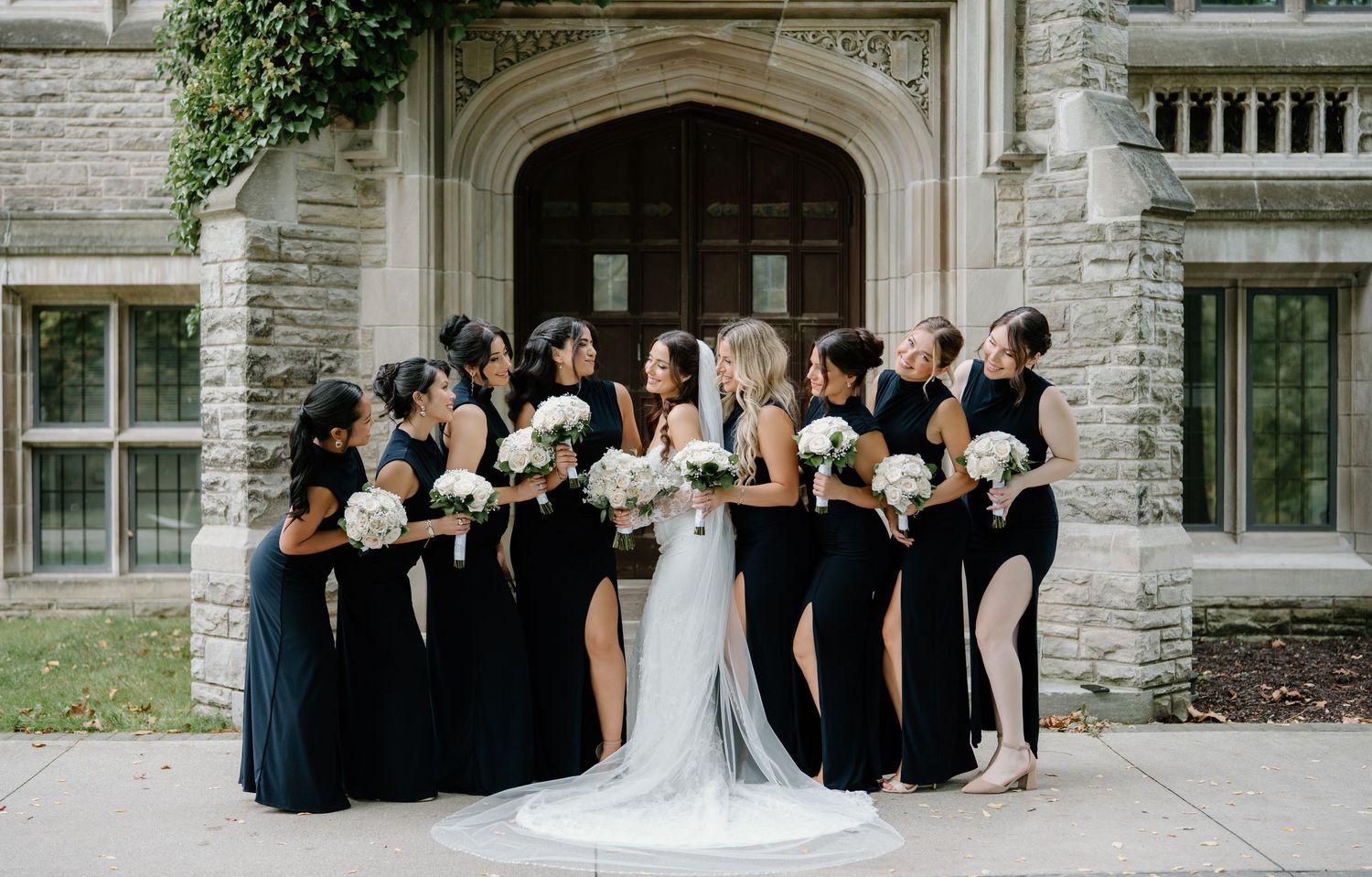 Wedding party members in navy dresses and formal attire sit together on stone steps of a historic building.