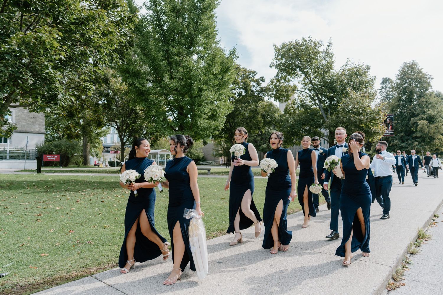 Wedding party walks in a line along a tree-lined pathway during an outdoor summer celebration.