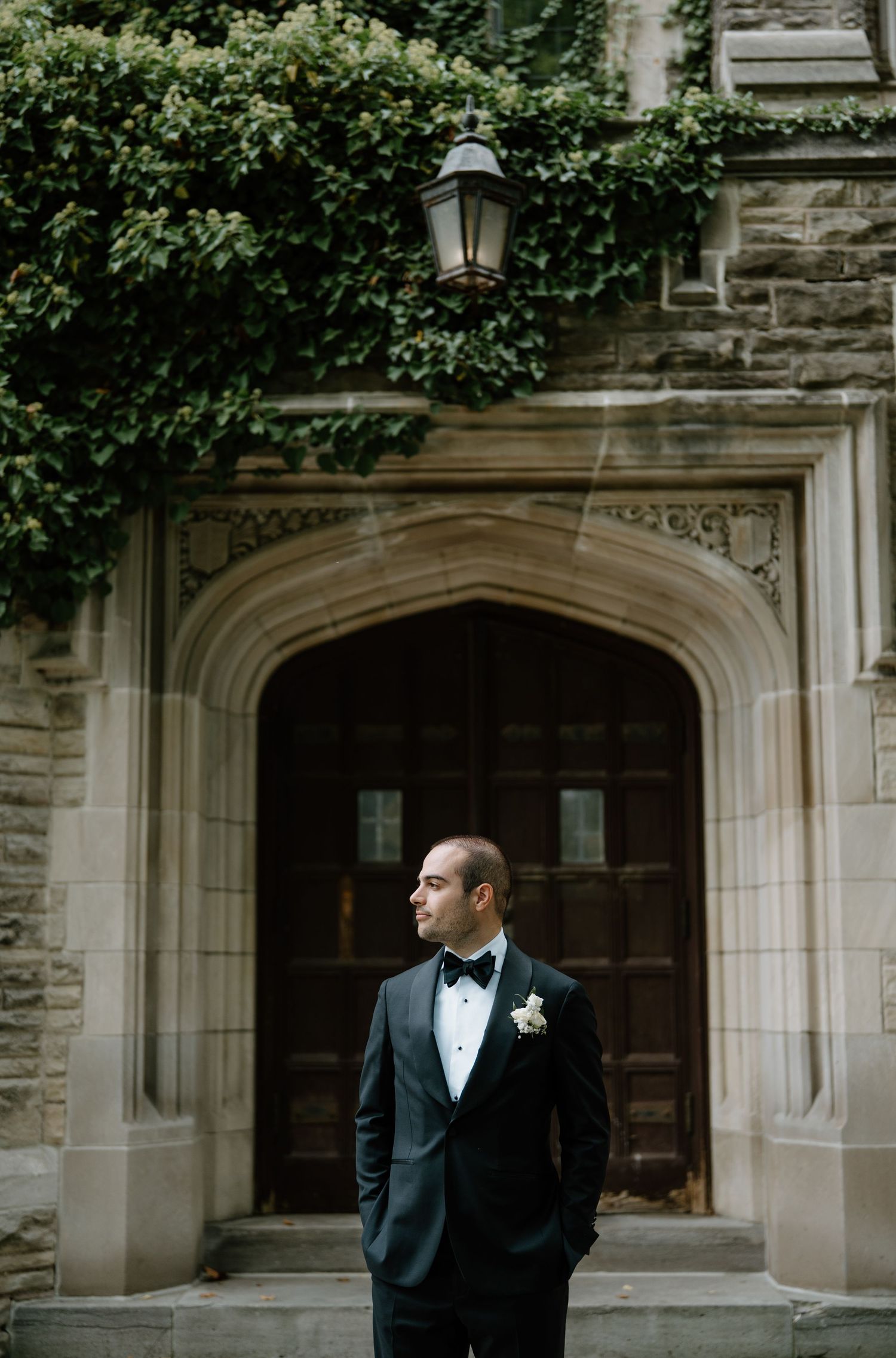 A black-tie wedding portrait against a stone archway with lush green ivy and classic architecture.