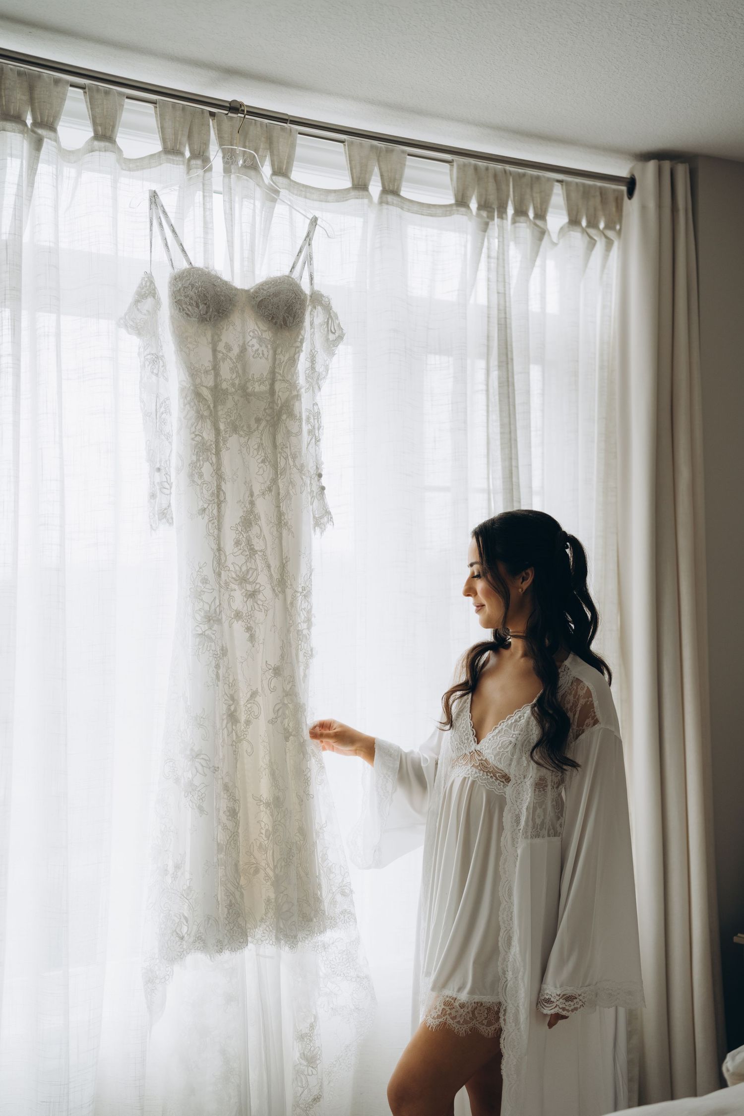 A person in a white robe stands near sheer curtained windows in soft morning light.