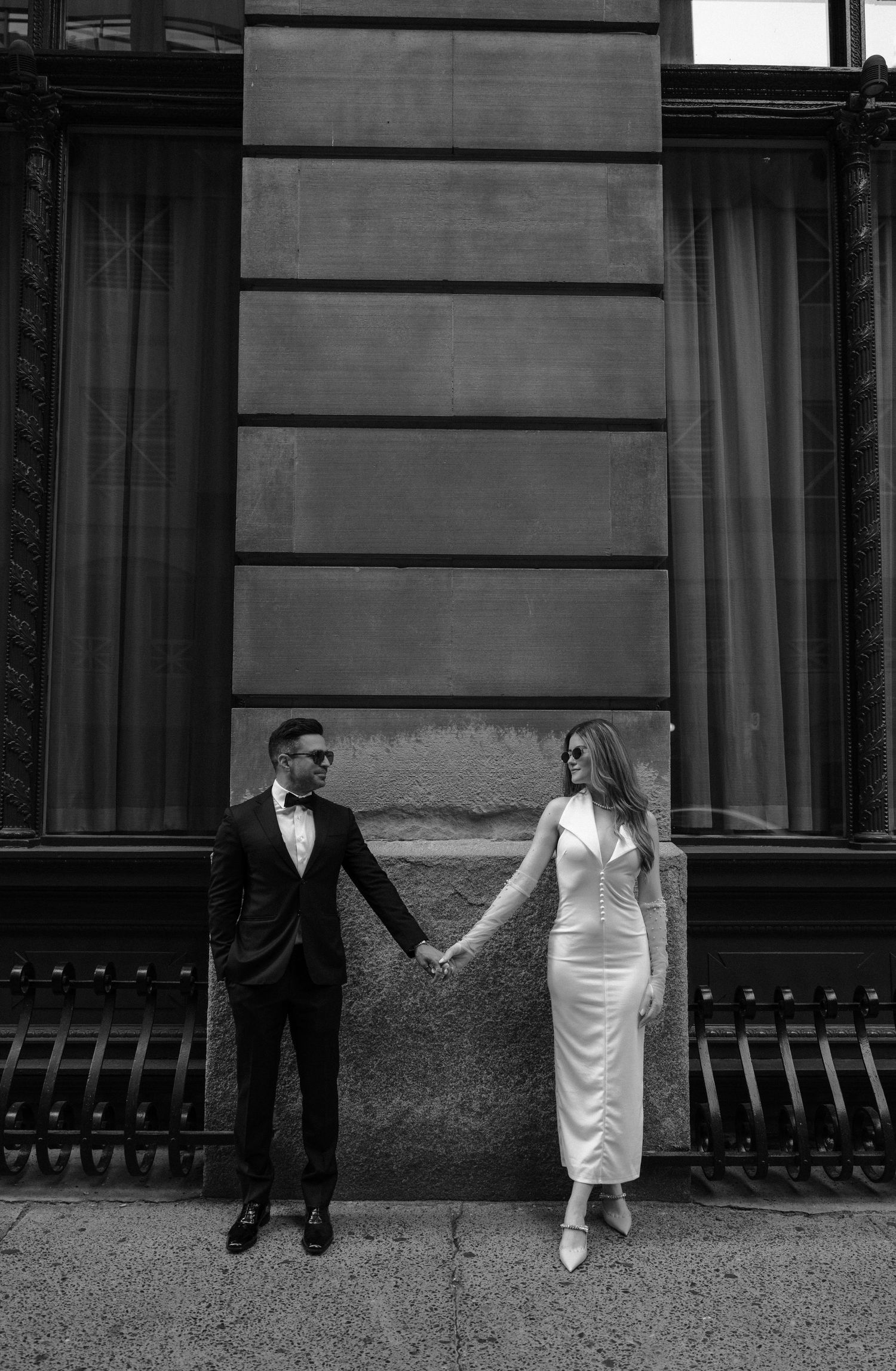 A couple in formal attire holds hands against an urban building wall in black and white photography.
