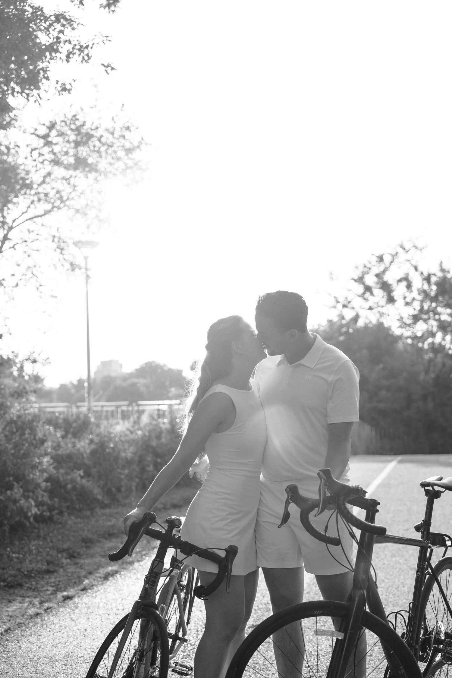 A romantic couple in white outfits share a tender moment while standing with their bikes on a sunny path.