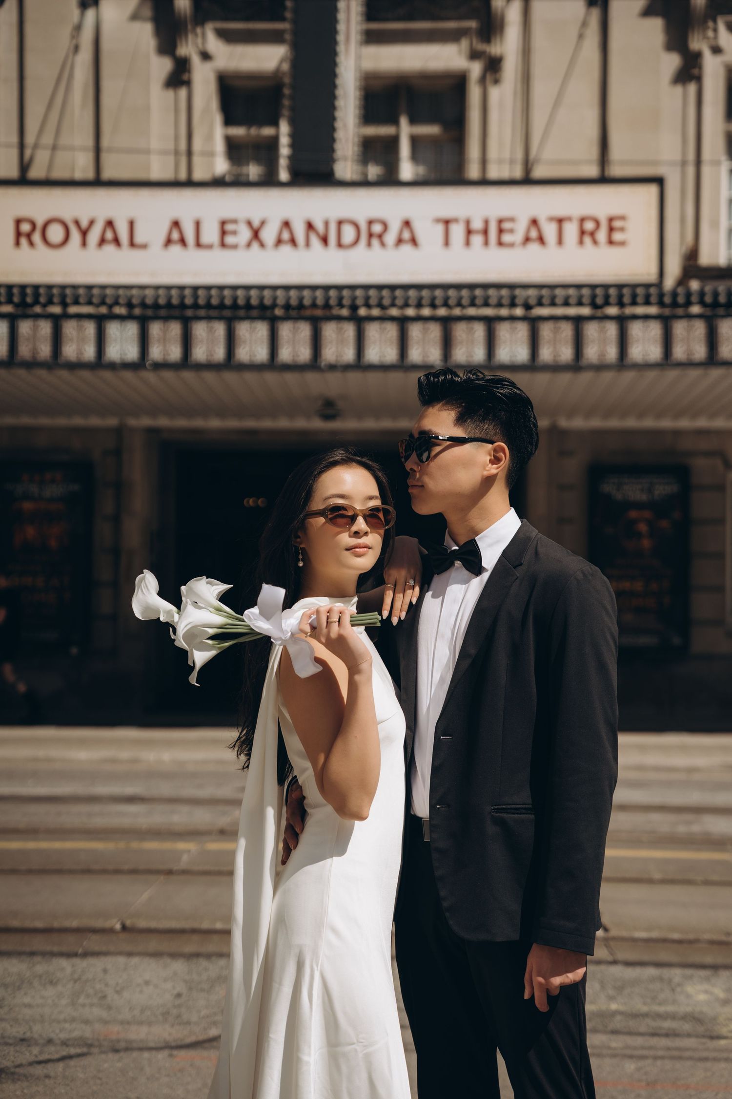 A stylish couple in formal wedding attire poses outside the Royal Alexandra Theatre in a romantic photoshoot.