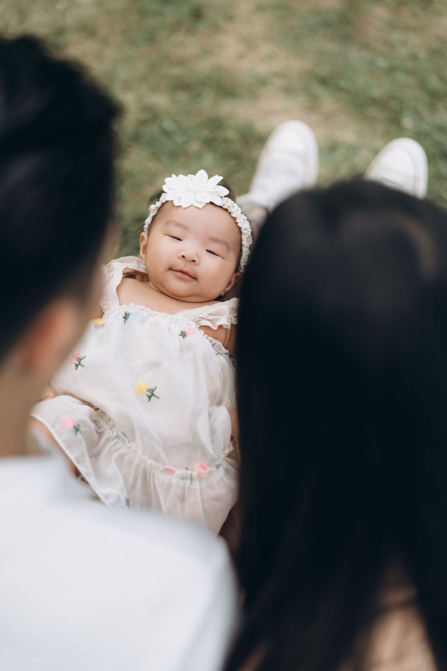 A baby wearing a delicate white dress with a floral headband smiles while being held.