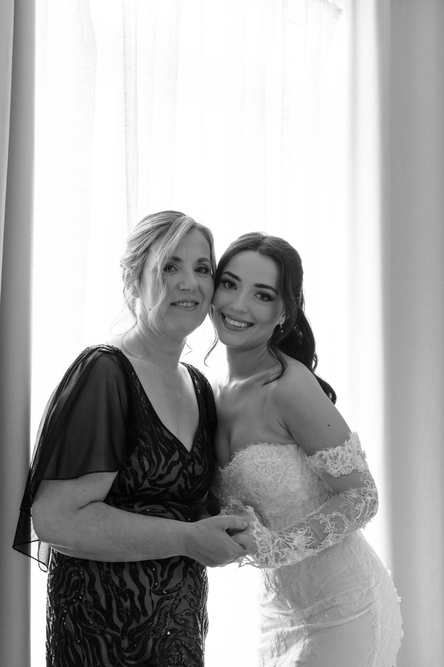 Black and white portrait of two women in formal attire standing together by a window in an intimate moment.