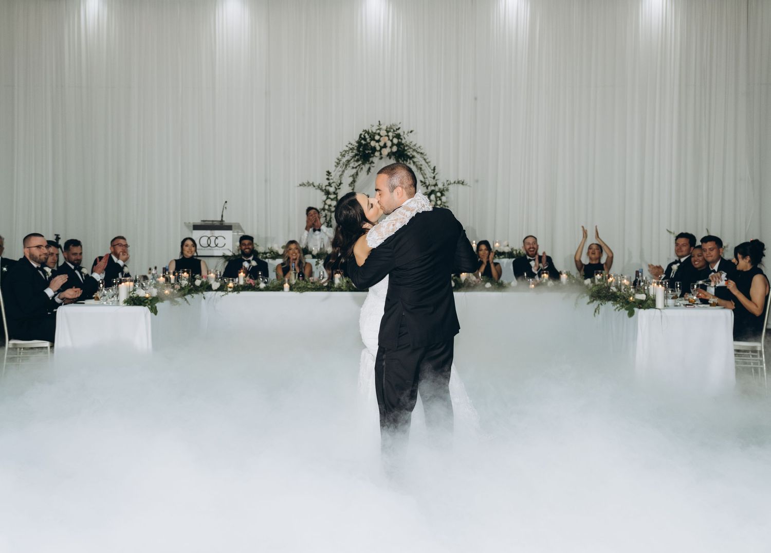 A wedding party dances together during a reception in a white-draped banquet hall.