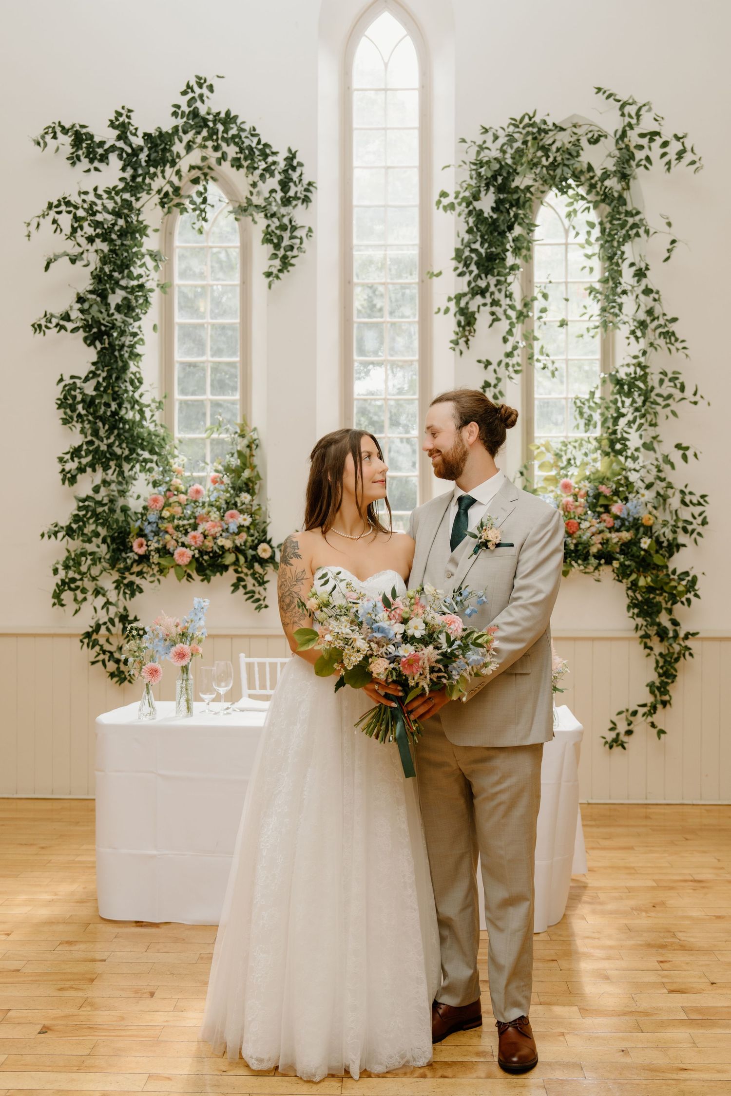 A couple shares a romantic moment in an elegant white venue decorated with climbing vines and floral arches.