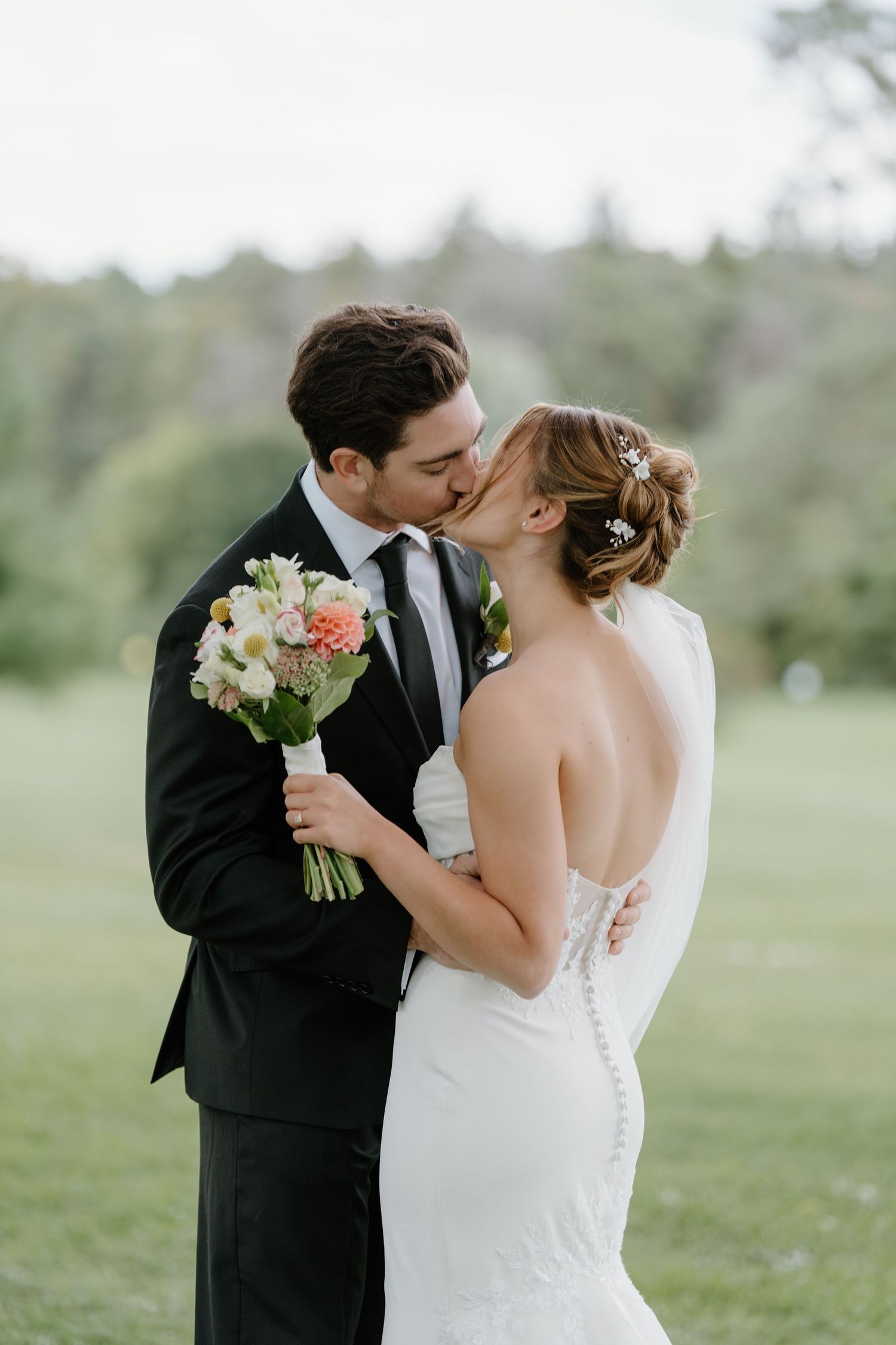 A romantic wedding couple shares a tender kiss while holding a bouquet of white and peach flowers in an outdoor setting.