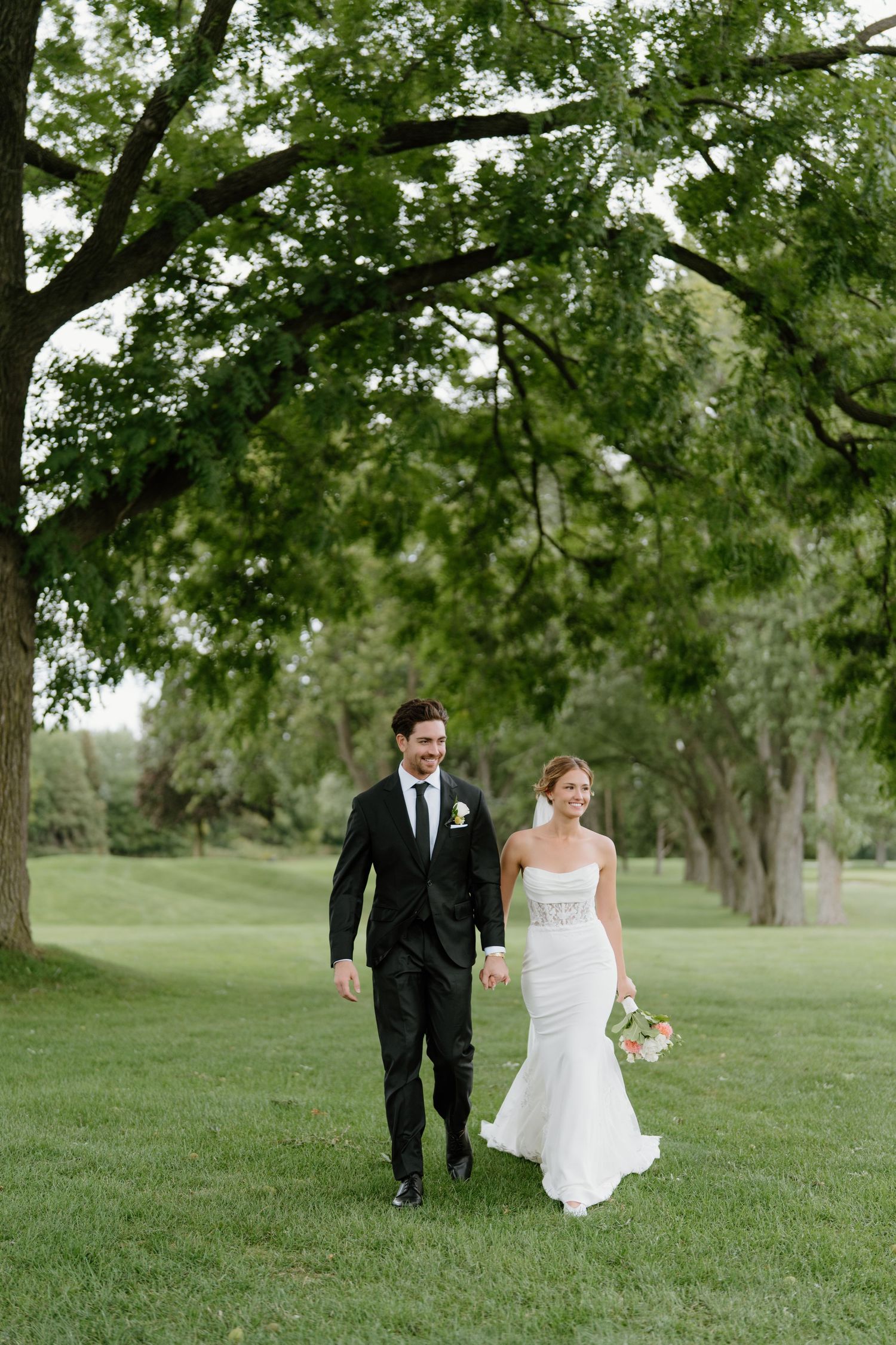 Newlyweds strolling across a scenic grassy field surrounded by mature trees.