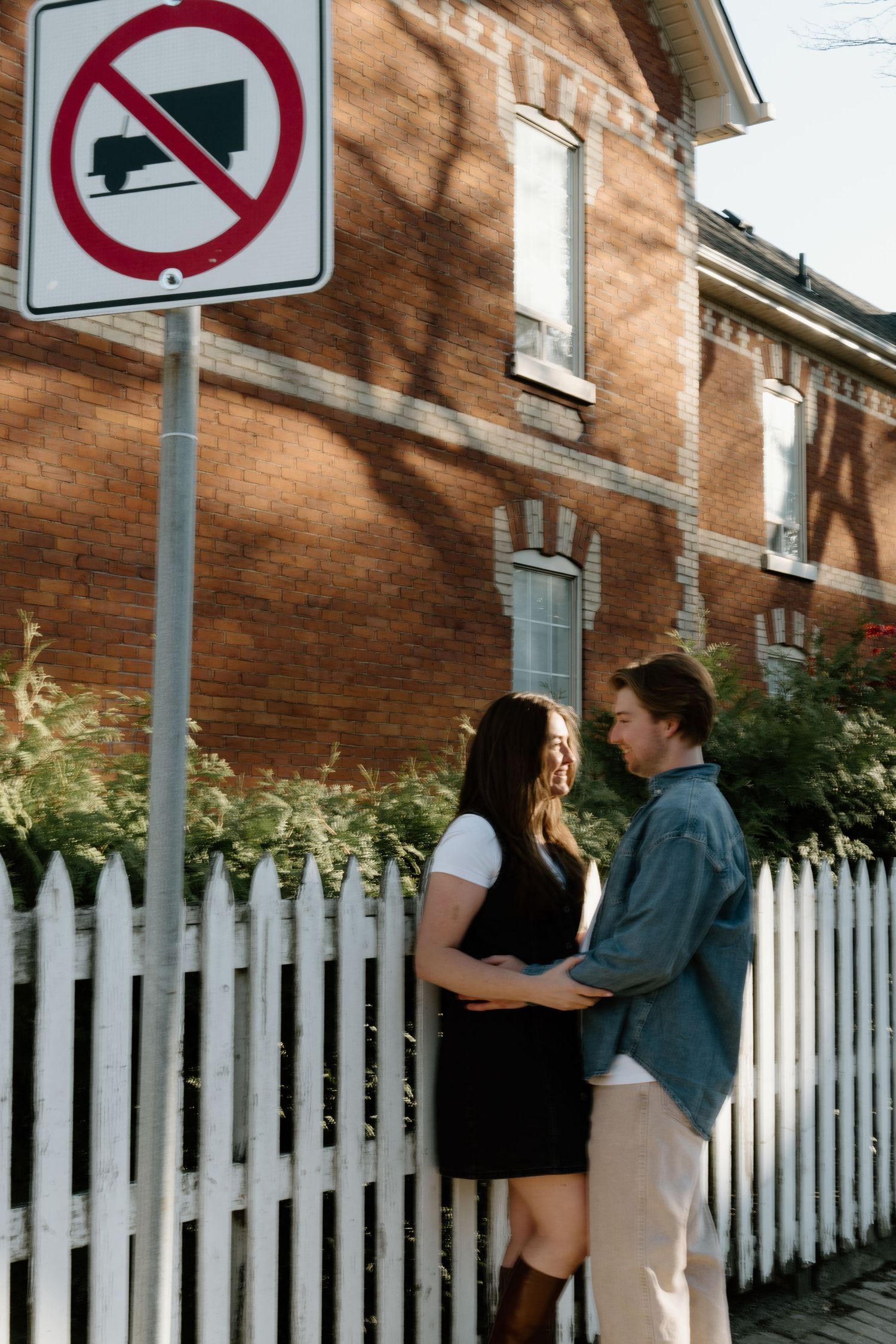Two people stand together near a vintage brick building with a white picket fence and a no trucks traffic sign.