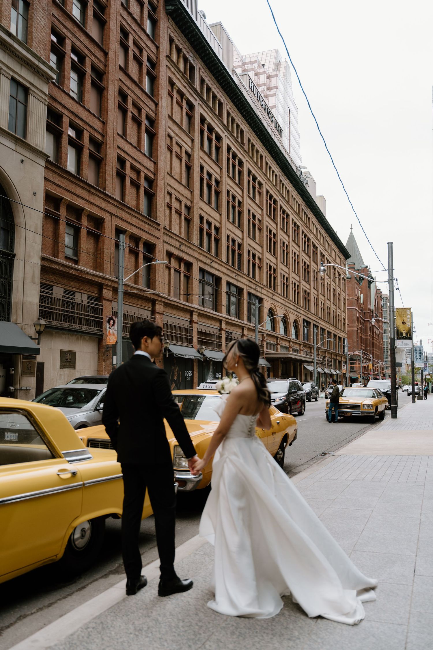 A couple in wedding attire walking past a historic brick building with yellow taxis on a city street.