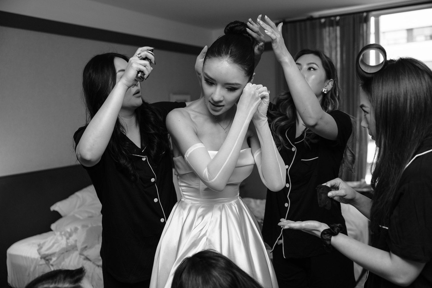 Someone in a white formal dress gets help with styling and preparations from multiple assistants in black attire backstage.