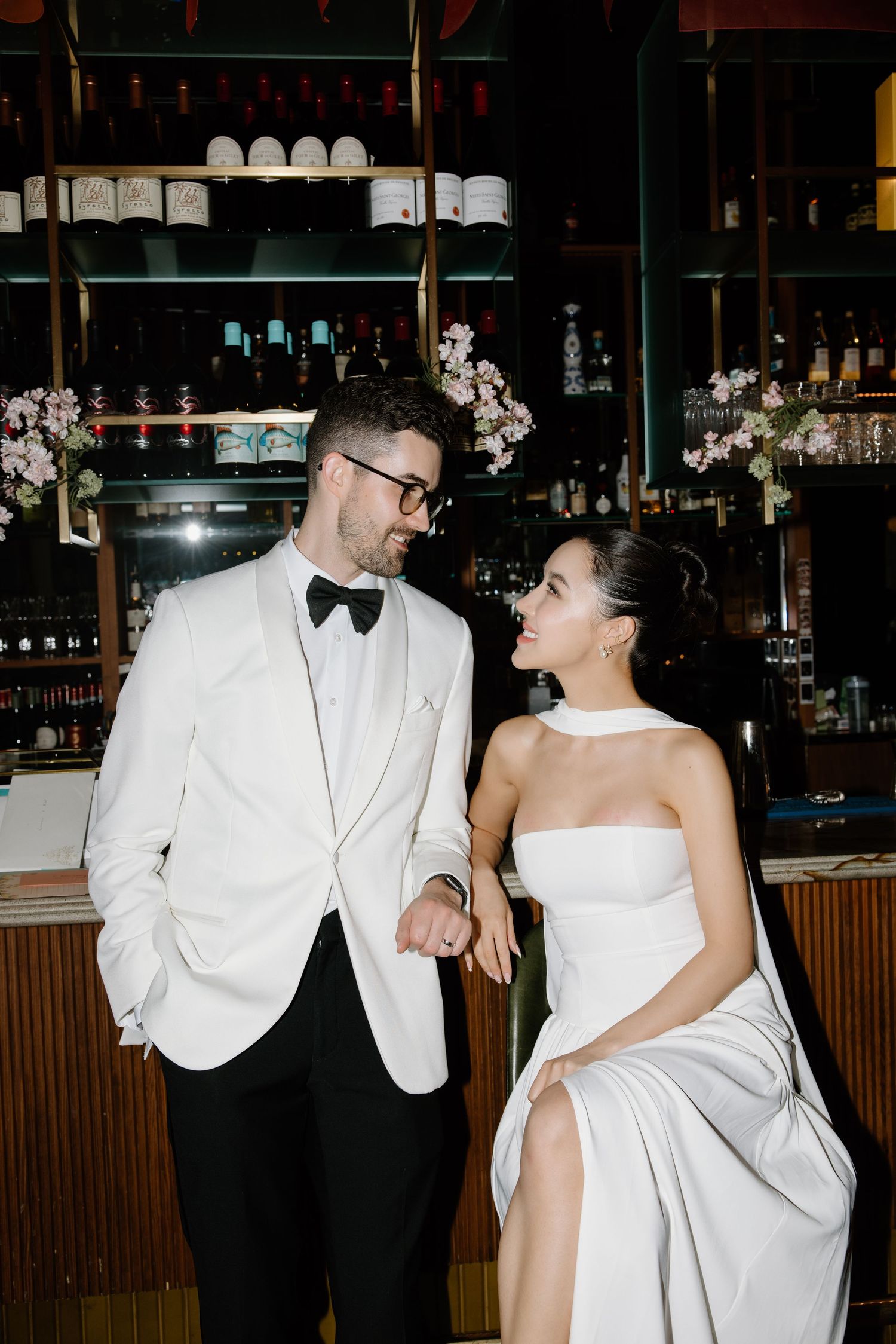 A couple in formal attire sharing a romantic moment at an elegant bar with shelves of bottles and glassware in the background.