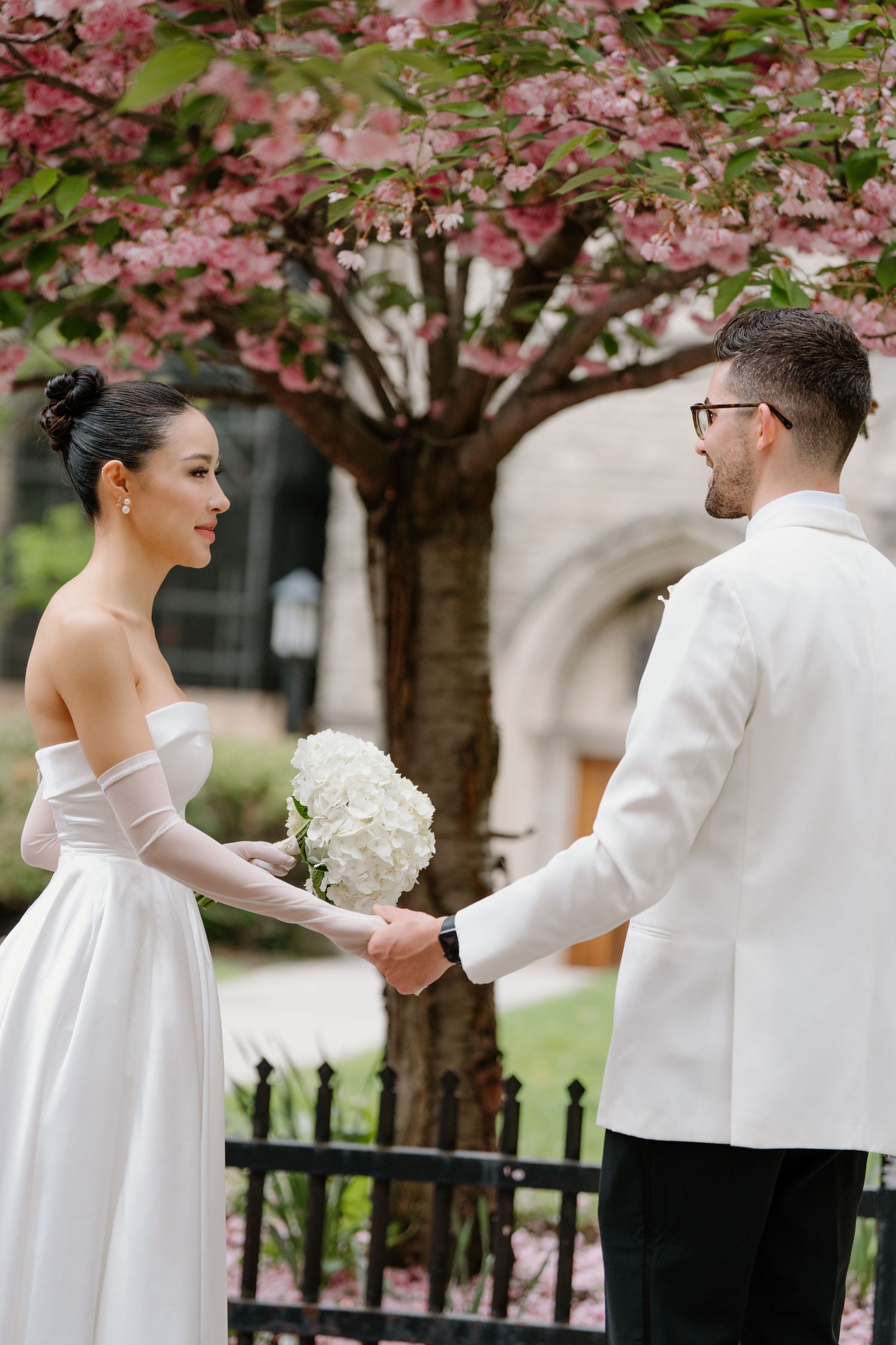 A couple holds hands under a blooming pink cherry blossom tree during a spring wedding ceremony.
