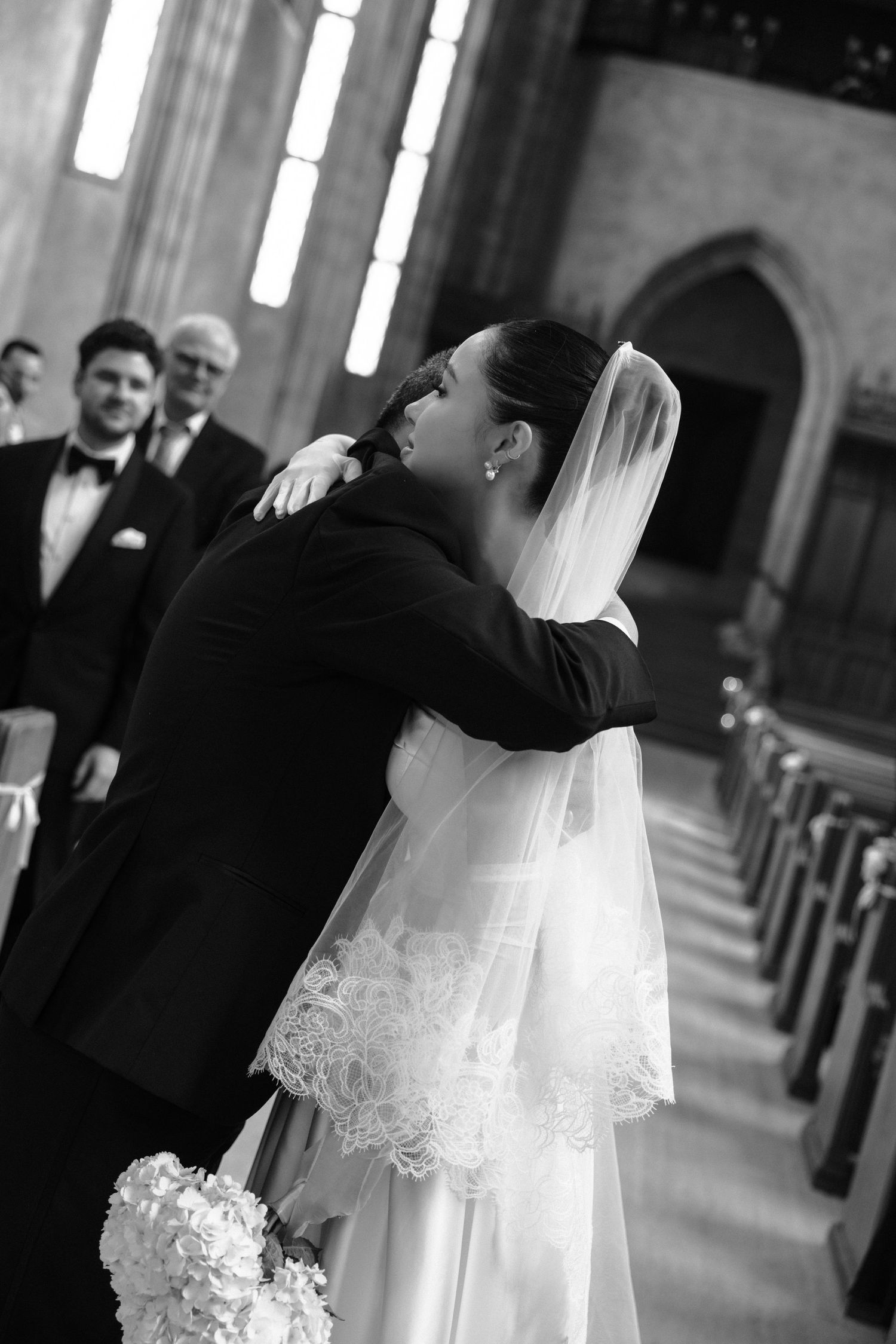 Black and white wedding photograph showing an emotional embrace during a church ceremony with arched architecture.