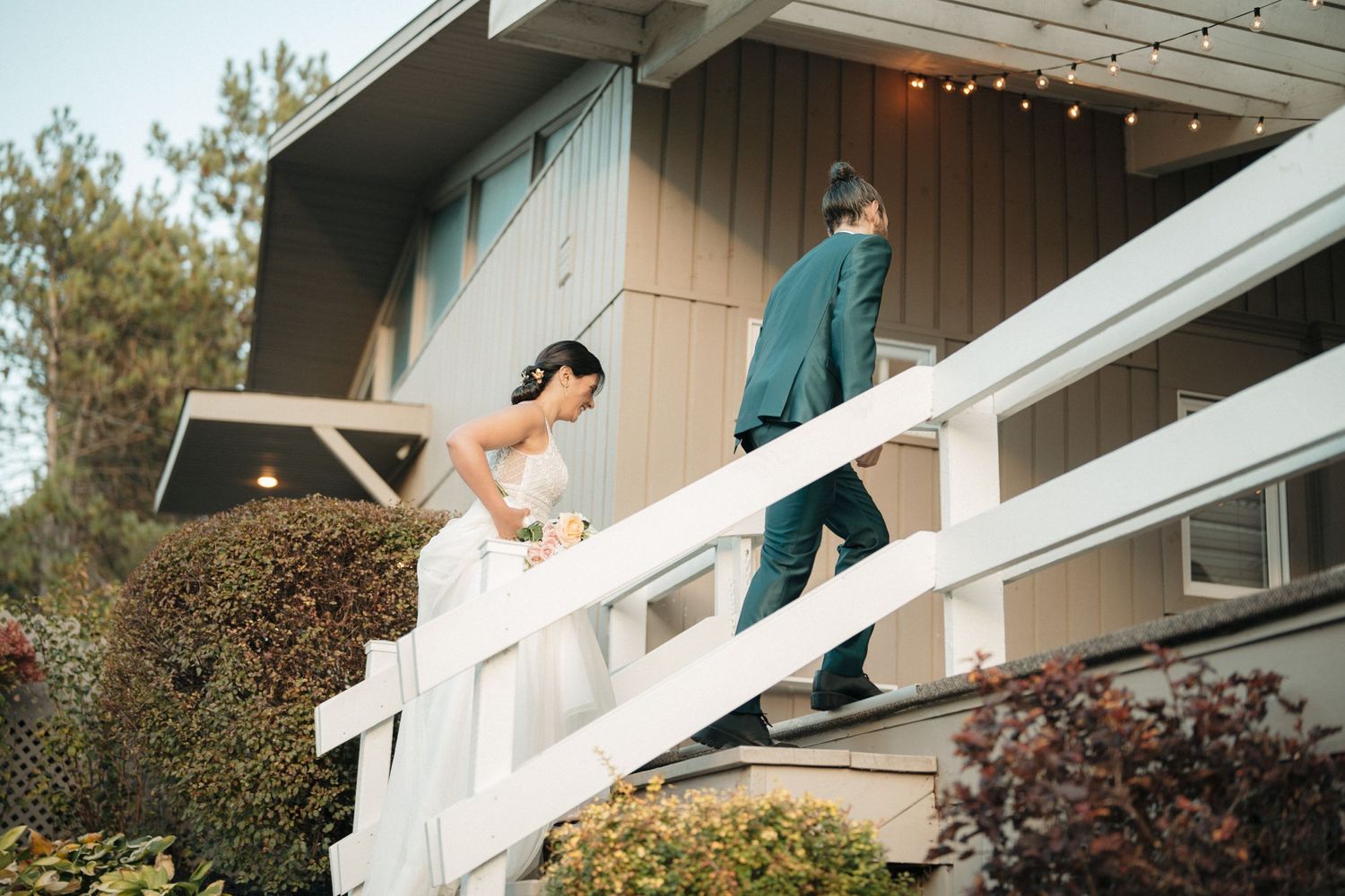 A bride in a white dress carefully walks down exterior stairs with someone walking ahead of her.
