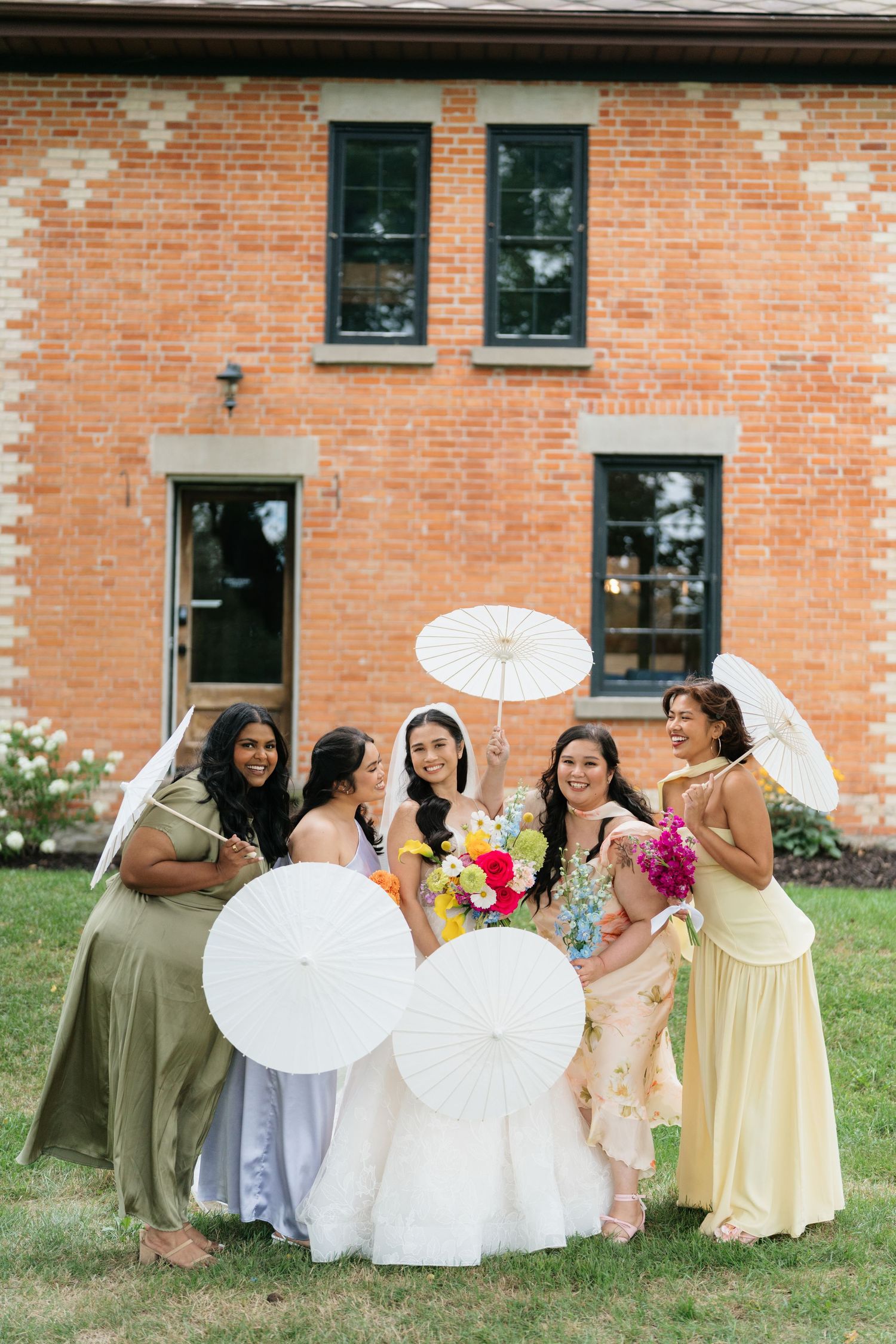 Wedding party members in pastel dresses hold parasols while posing in a line against a brick building exterior.