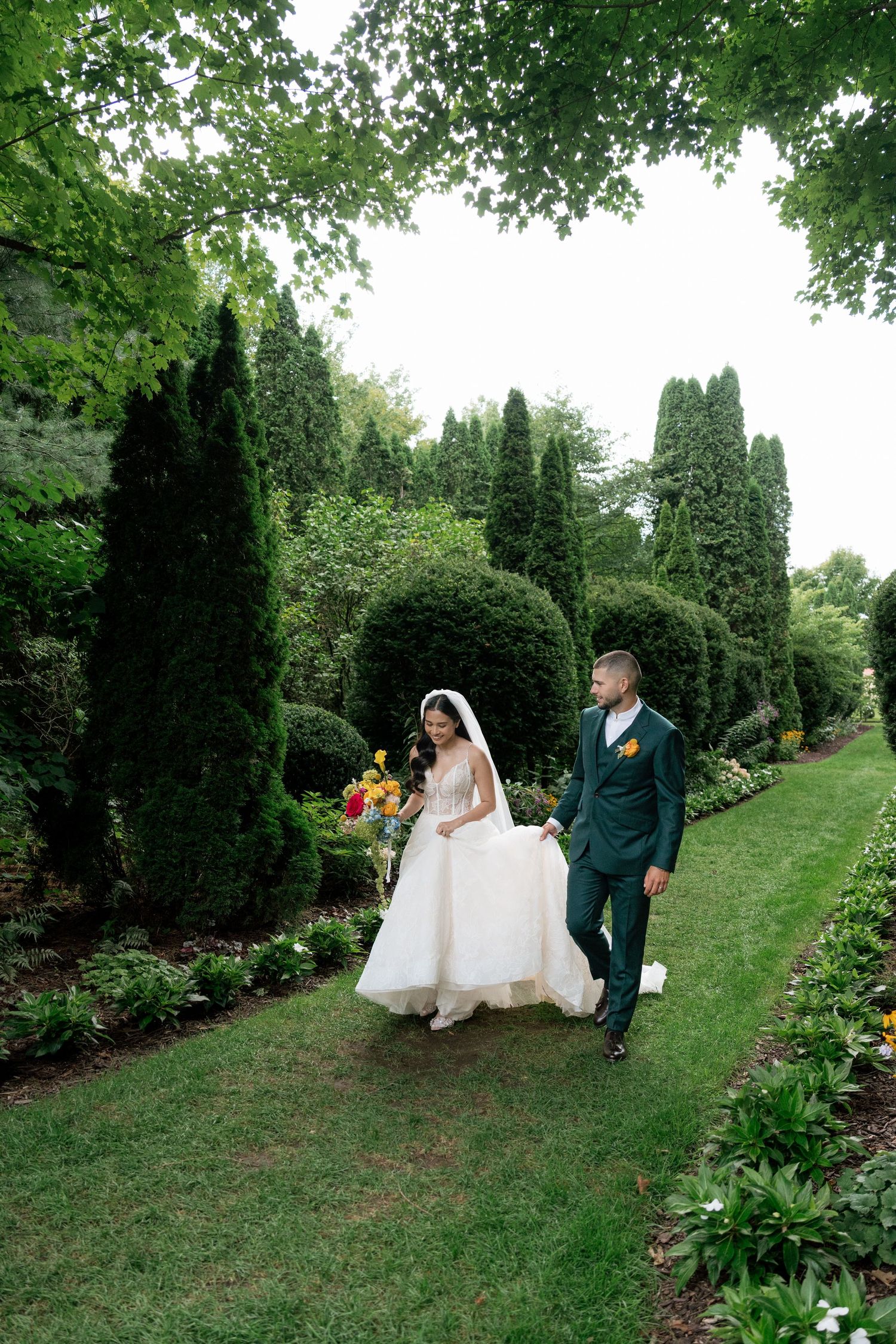 Wedding couple sits together on a grassy hillside surrounded by evergreen trees in a lush garden setting.