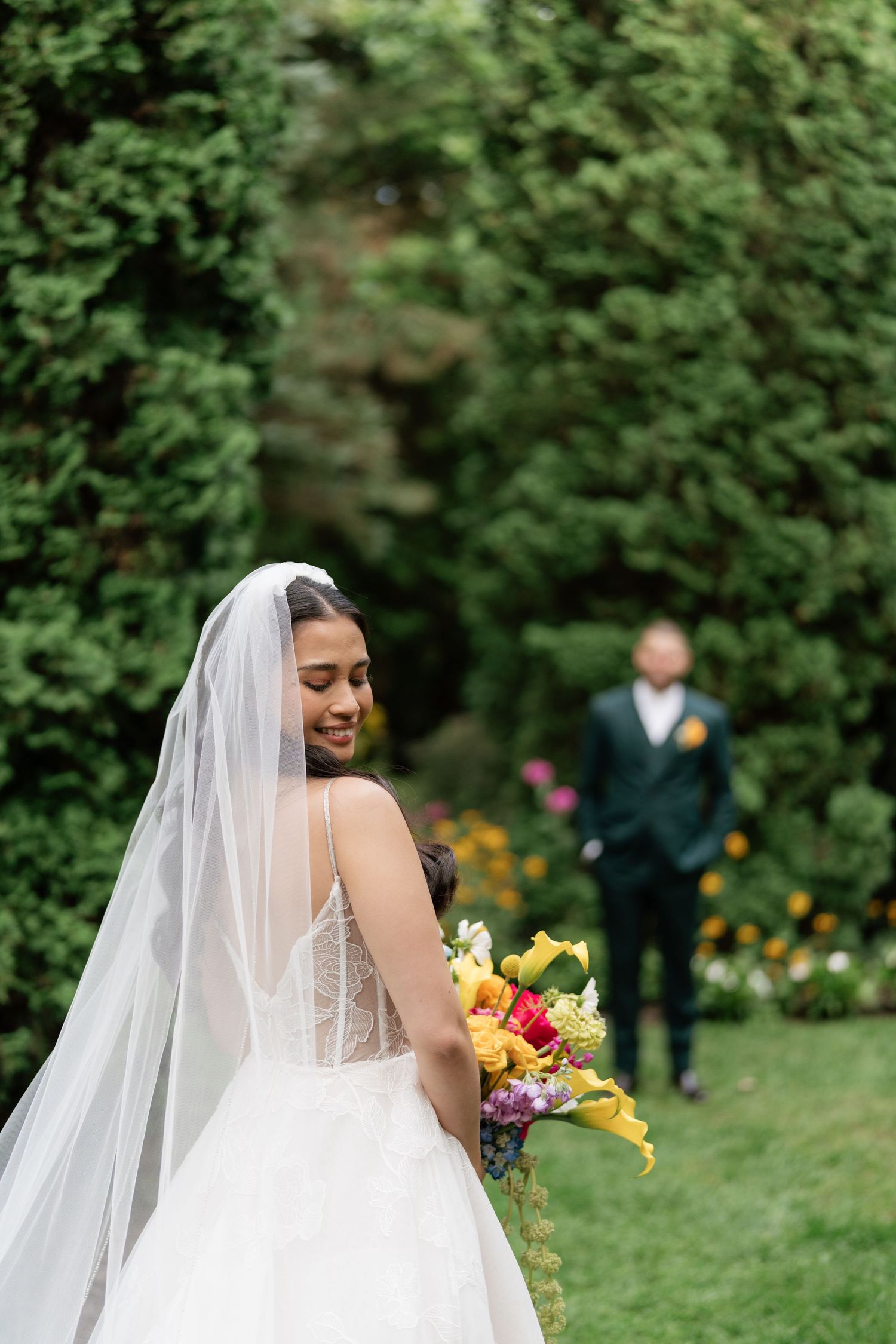A bride in a white wedding dress holds a vibrant yellow and pink bouquet in a lush garden setting.