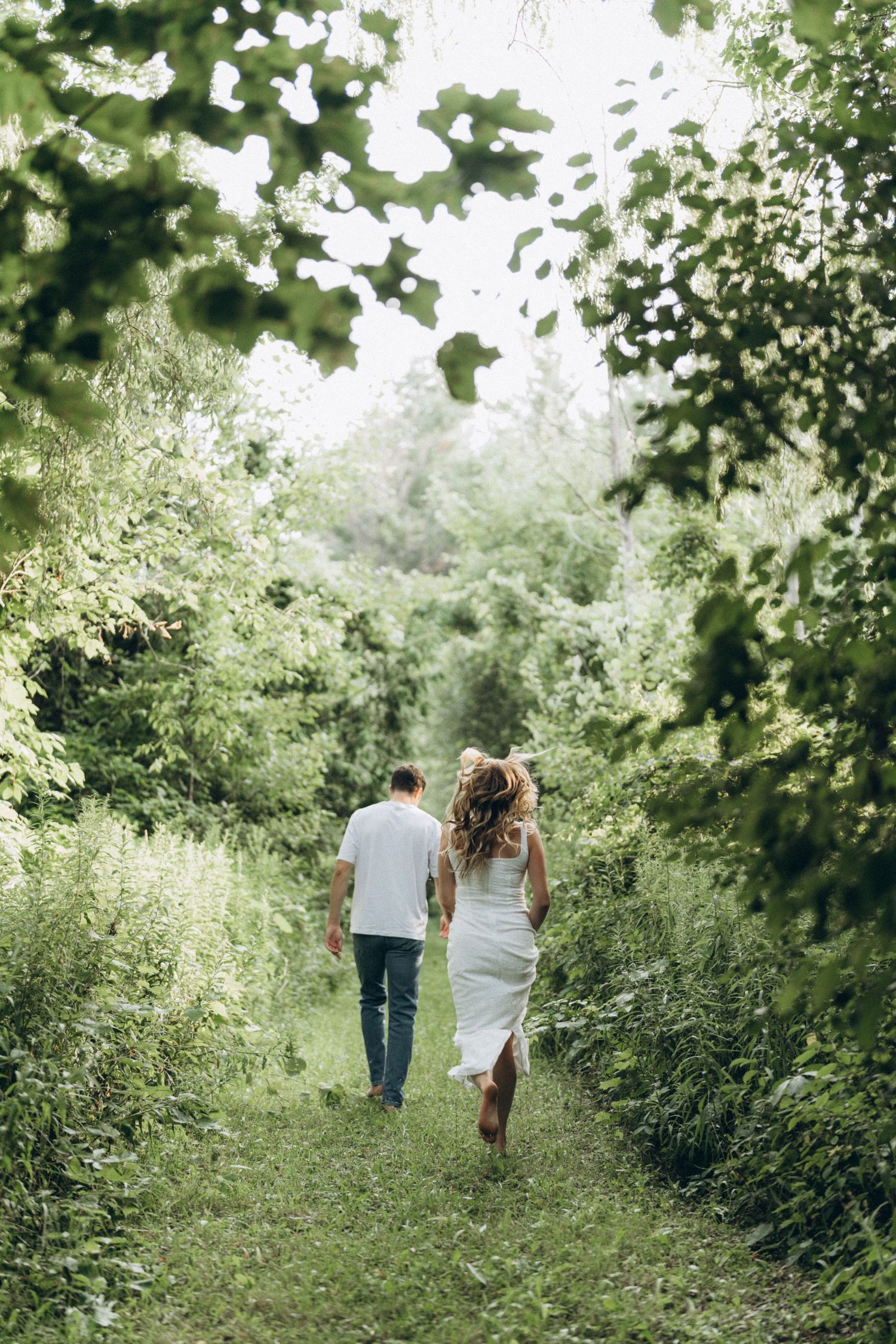 A couple walks hand in hand down a lush green nature path surrounded by trees and foliage on a bright summer day.