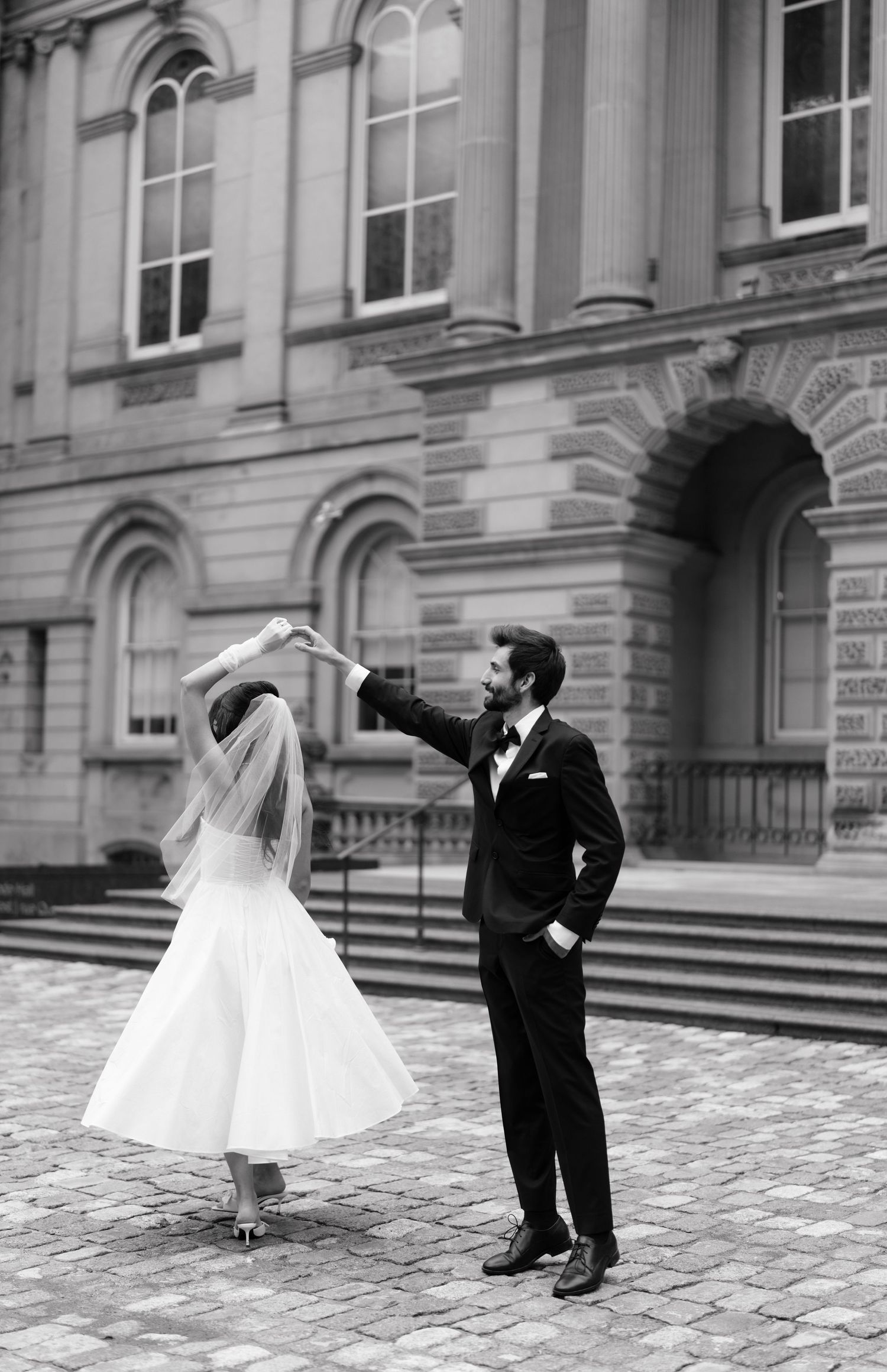 A sequence of black and white photos showing a couple in wedding attire dancing playfully on a cobblestone courtyard.