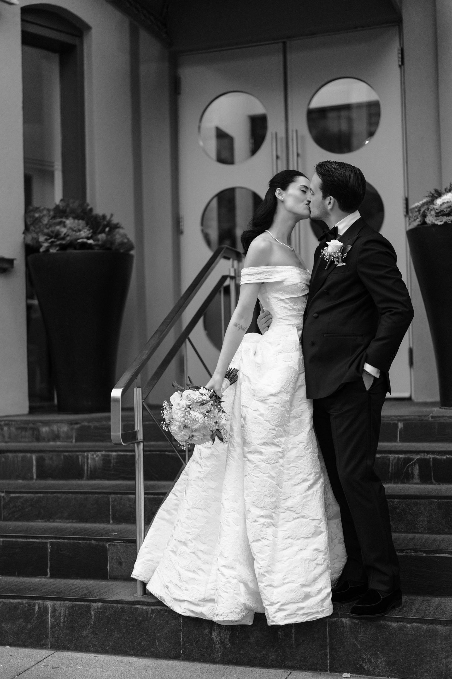 Black and white wedding photo showing bride and groom leaning against railing in romantic pose outside venue entrance.