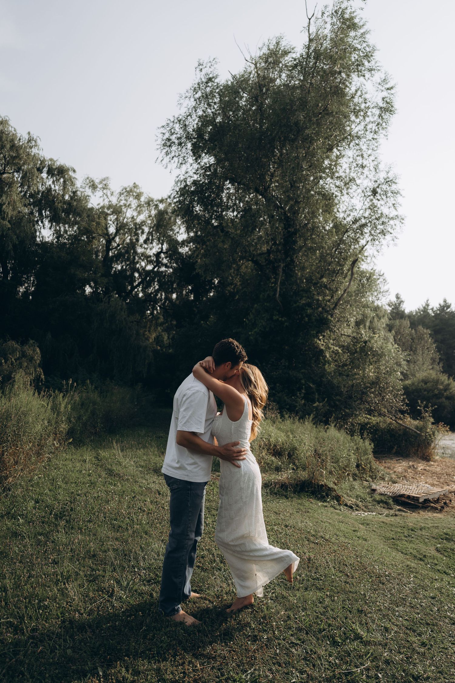 A romantic couple in wedding attire embrace while standing on a grassy hillside in a natural outdoor setting.