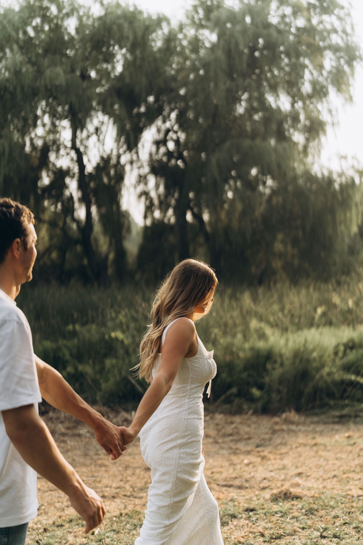 A couple in white outfits holds hands while walking through a sunlit field with willow trees in the background.