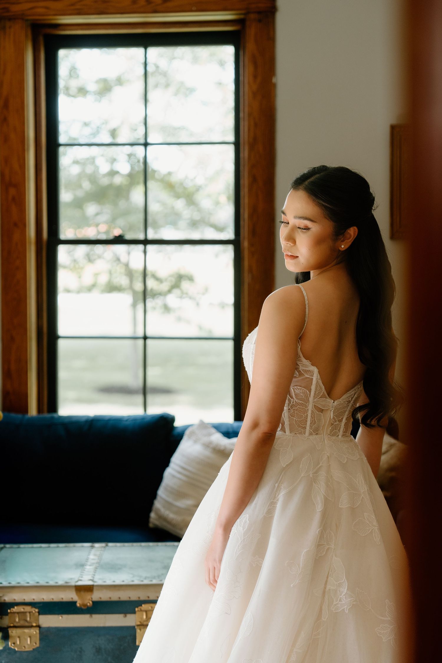 A bride in a flowing white gown sits on a rustic wooden bench near large windows in a natural light setting.