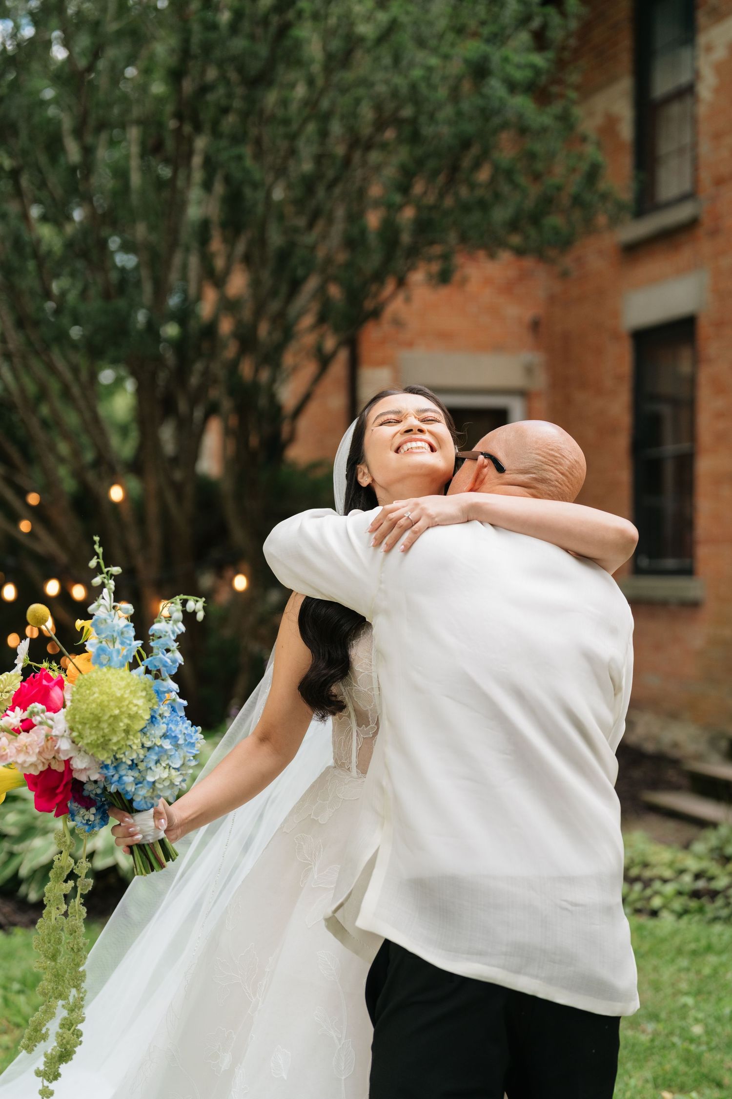 A joyful couple embraces outdoors in a garden setting with colorful flowers and a brick building in the background.