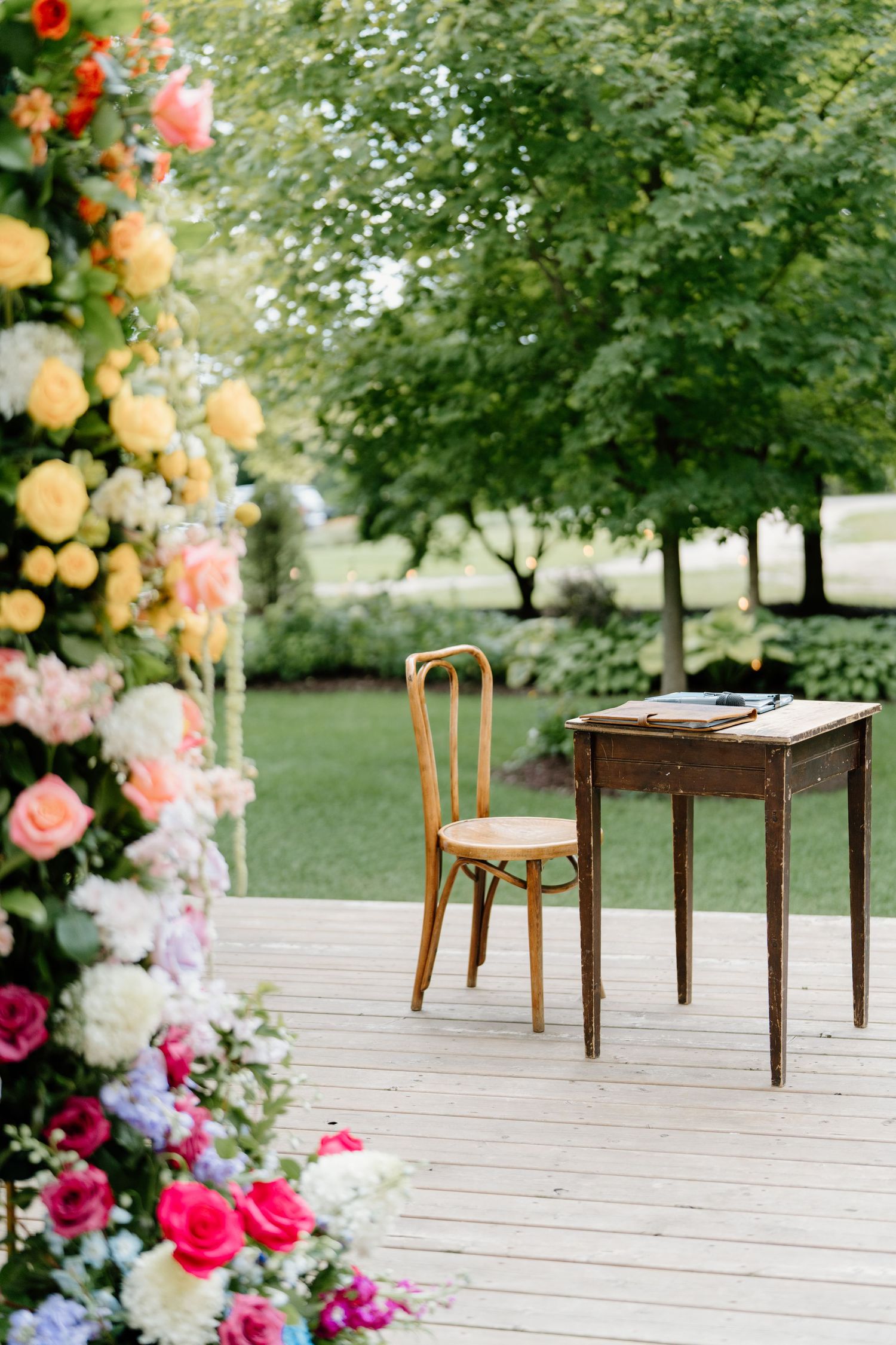Wooden chairs arranged on a white deck with colorful floral arrangements and greenery creating an outdoor wedding setting.